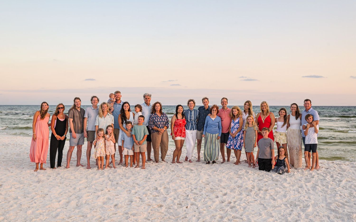 Family of eight poses together on a sandy beach at sunset, dressed in coordinating neutral and white outfits.