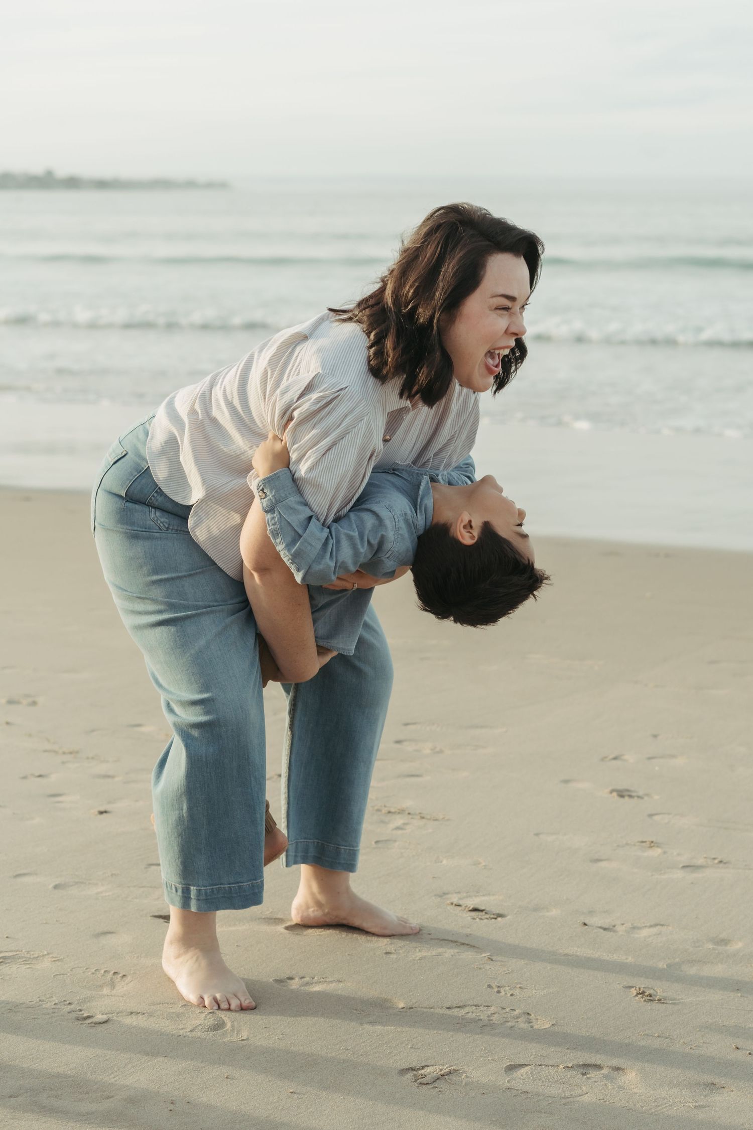 Two women laughing and playing on a sandy beach, one lifting the other near ocean waves.