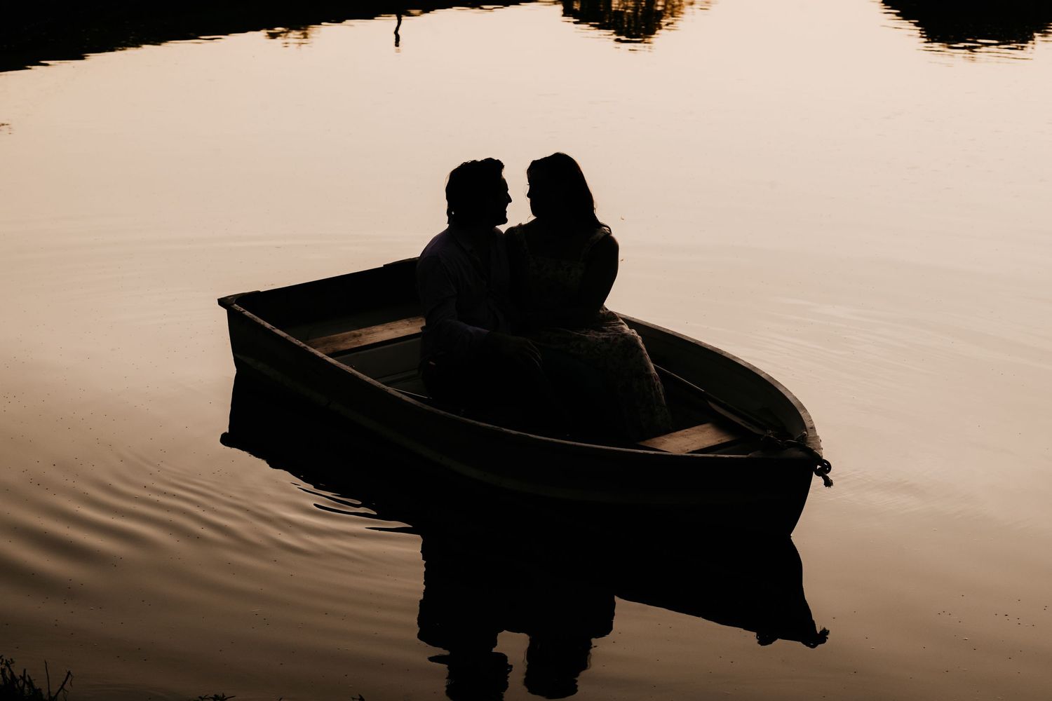 Silhouette of a romantic couple sitting face to face in a rowboat on a calm lake at golden sunset.