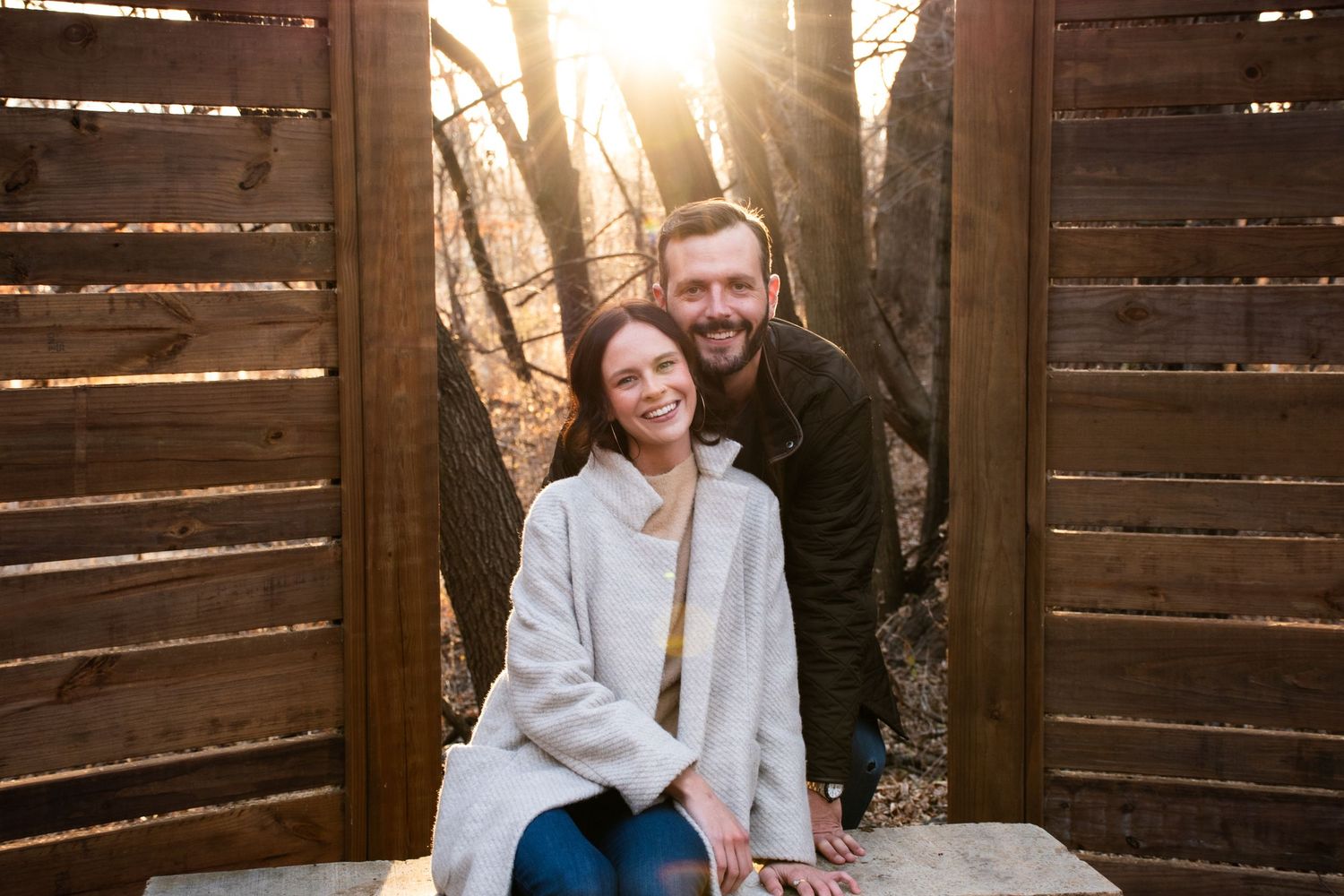 Smiling couple sitting together in a wooden gazebo with golden sunset light filtering through autumn trees.