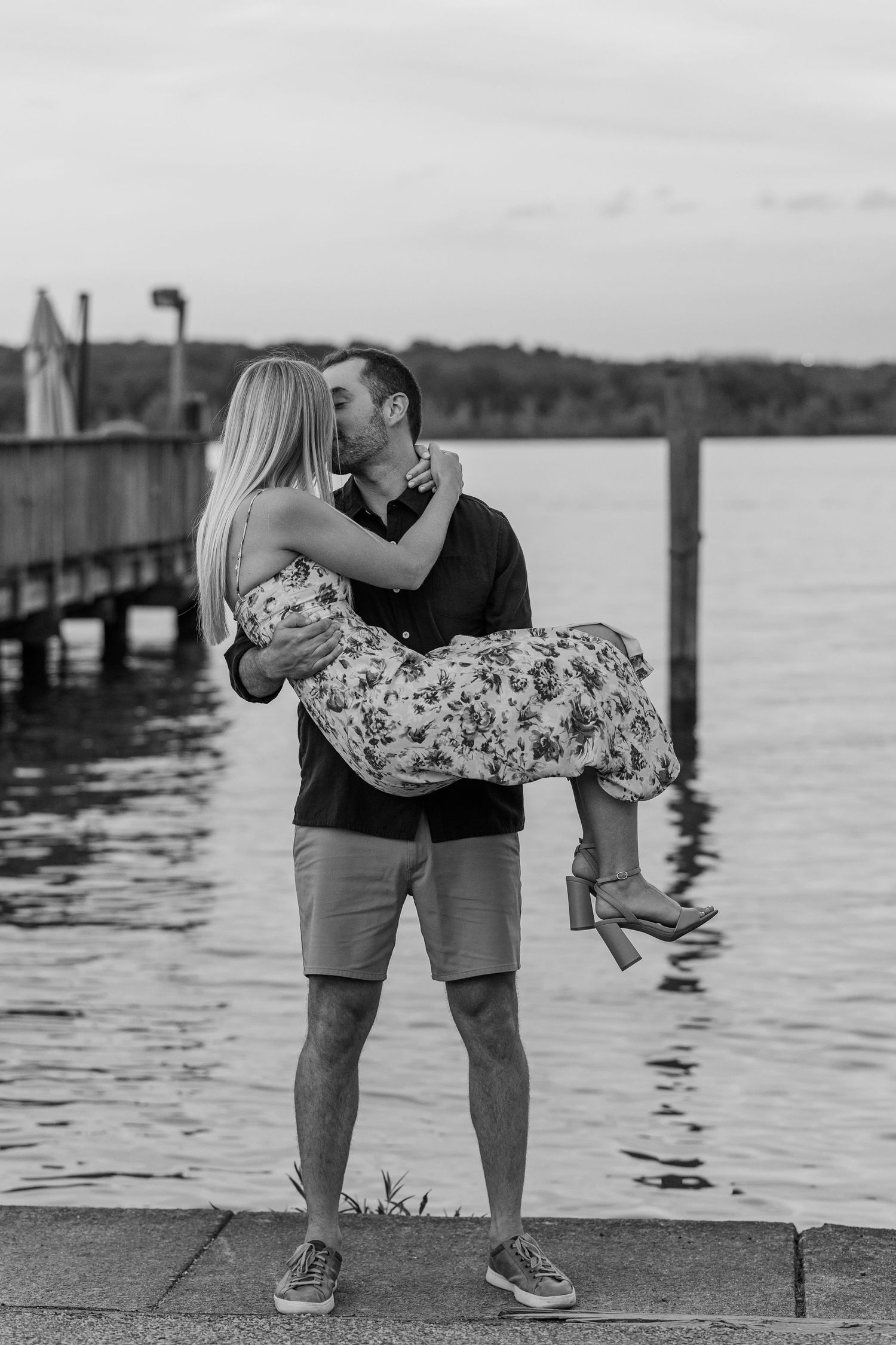 Black and white photo of a man carrying a woman in his arms while kissing near a waterfront dock at dusk.