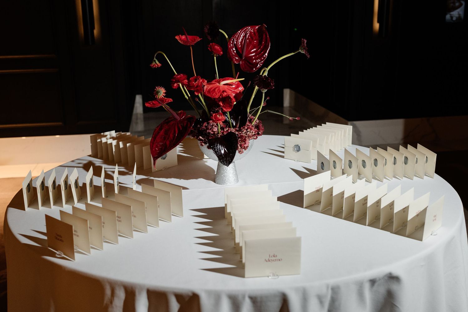 Elegant wedding escort card table with white place cards and red anthurium floral centerpiece on white tablecloth.