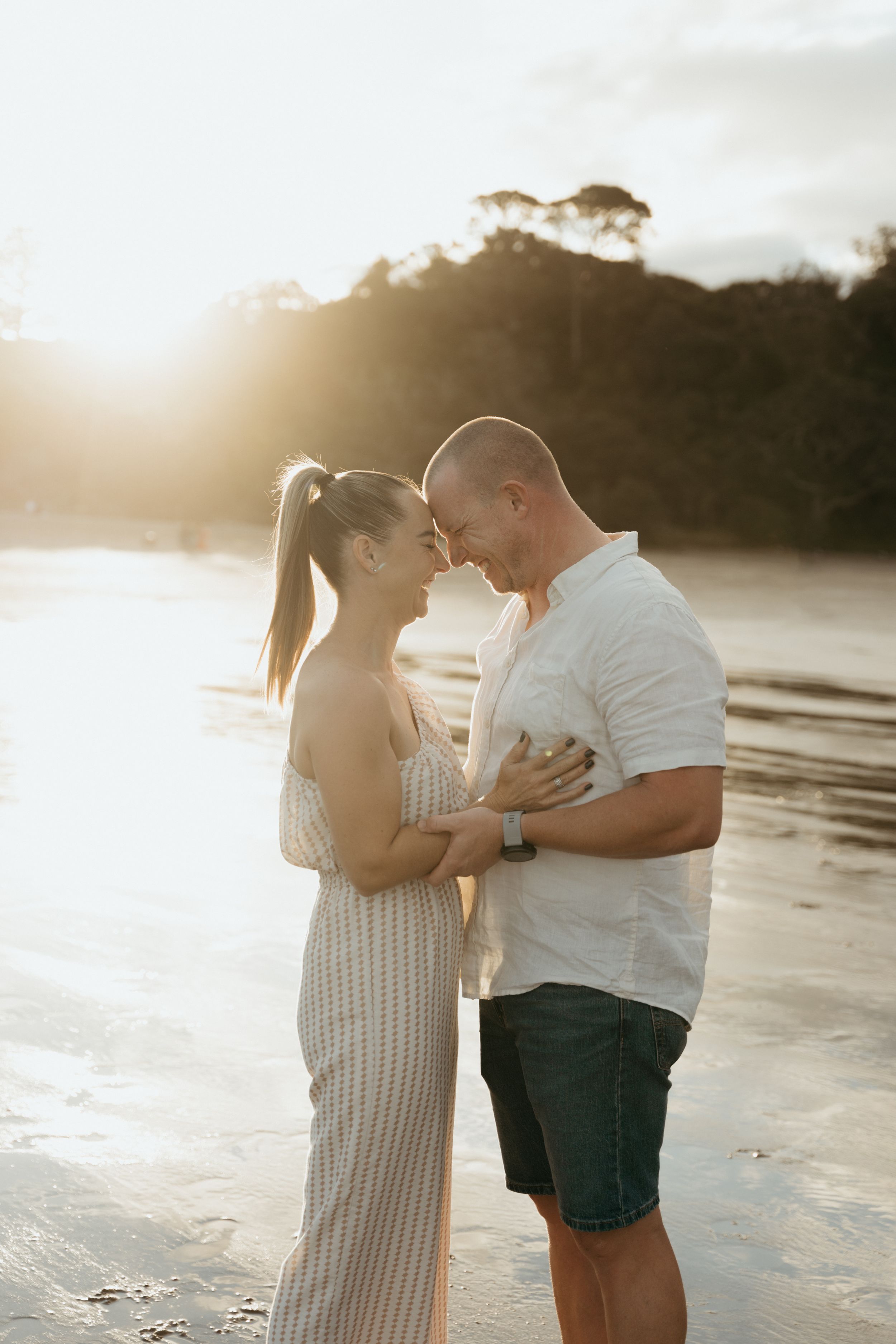Couple embracing on a sunlit beach at golden hour, smiling at each other in a romantic outdoor portrait.