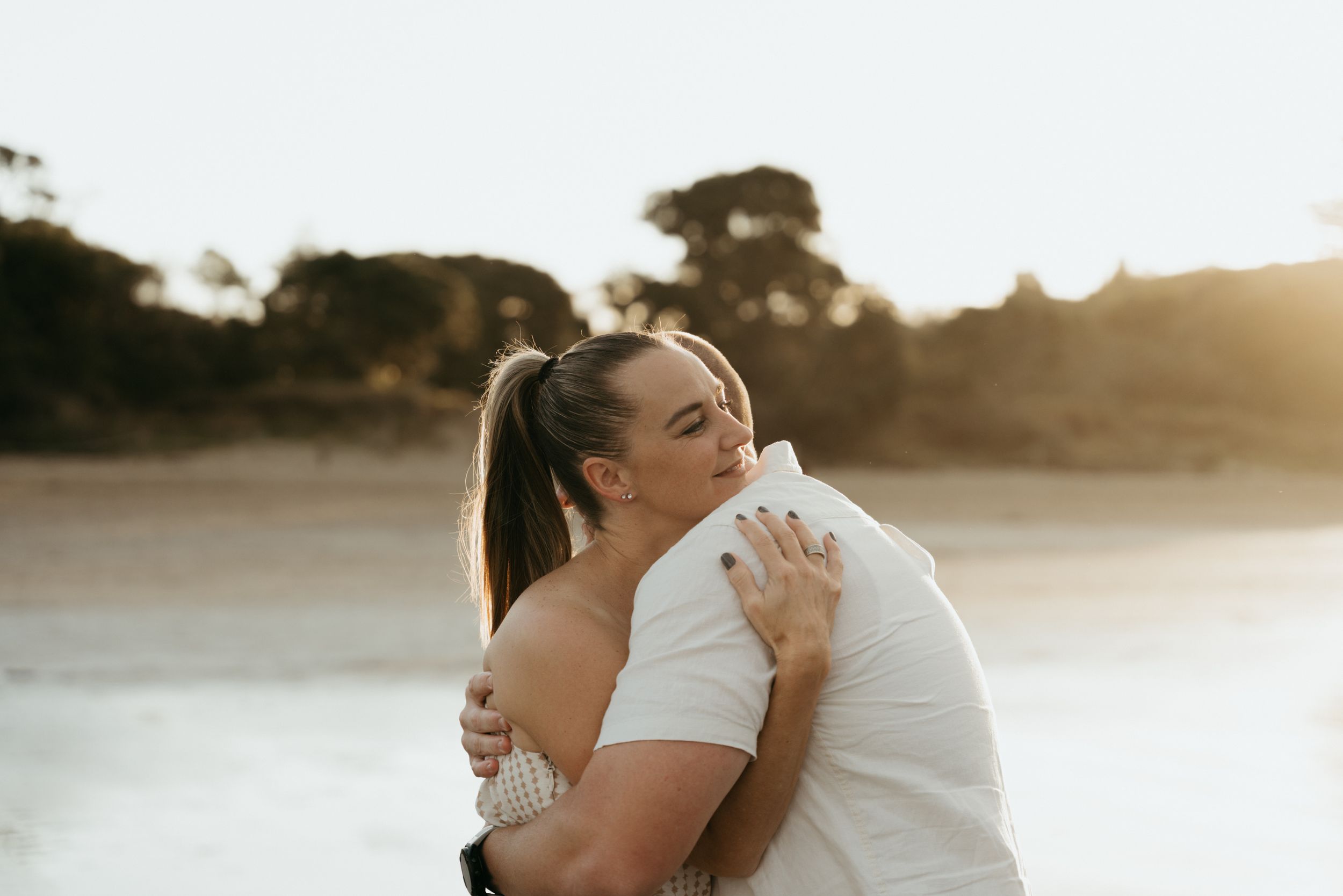 Couple embracing on a beach at golden hour with trees in the background.