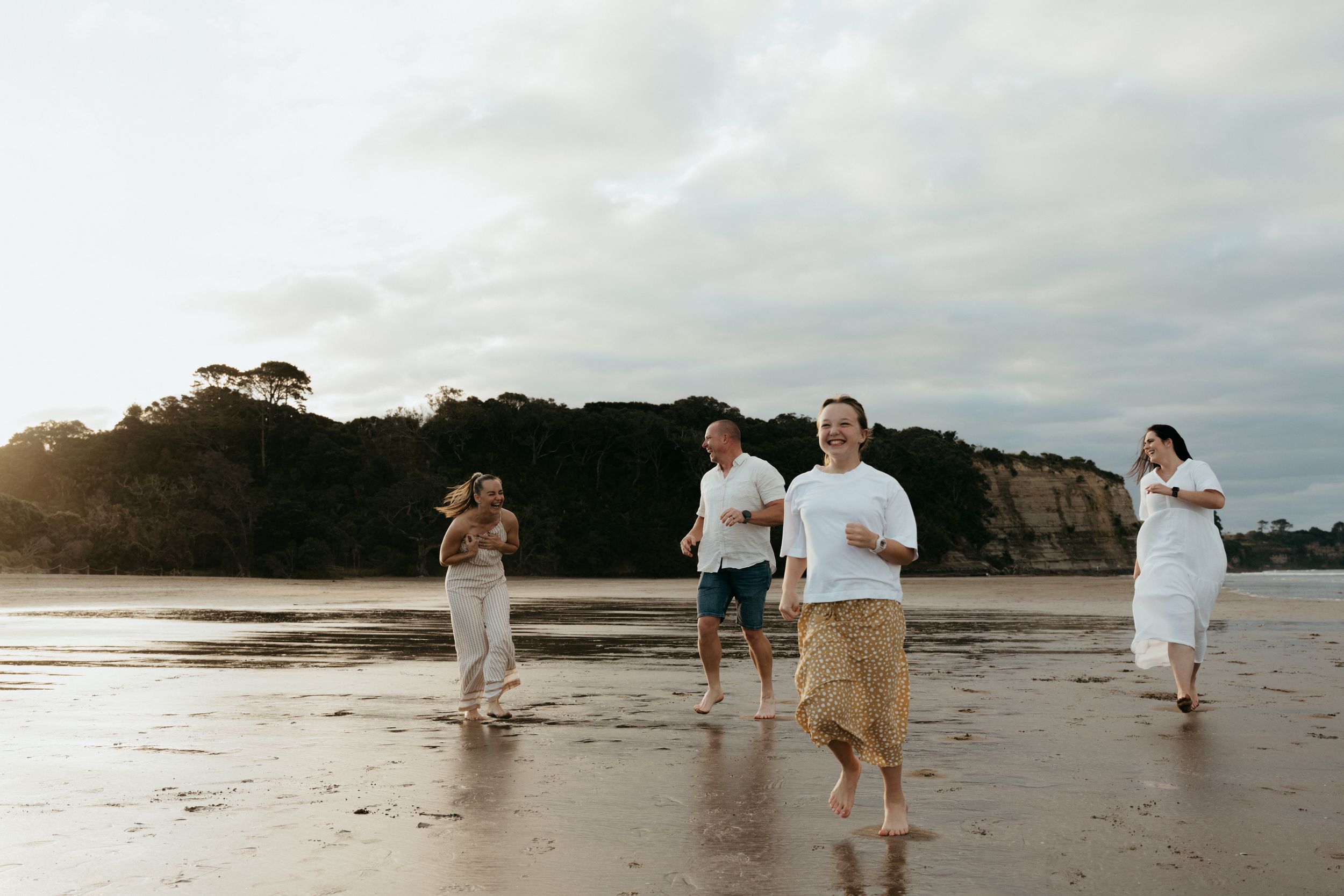 Group of friends running joyfully on a sandy beach at sunset with cliffs and trees in the background.