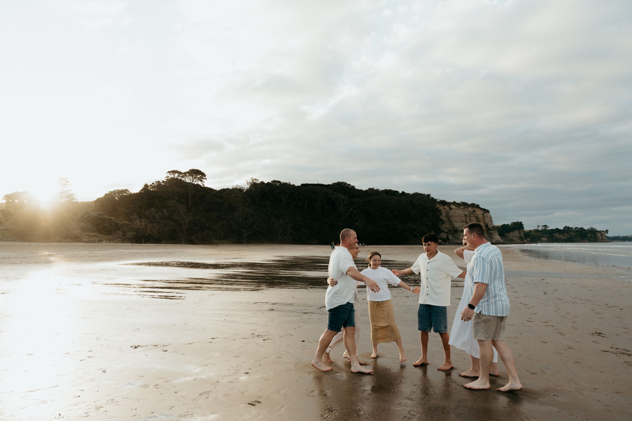 Family holding hands walking on a sandy beach at sunset with lush green hills in the background.