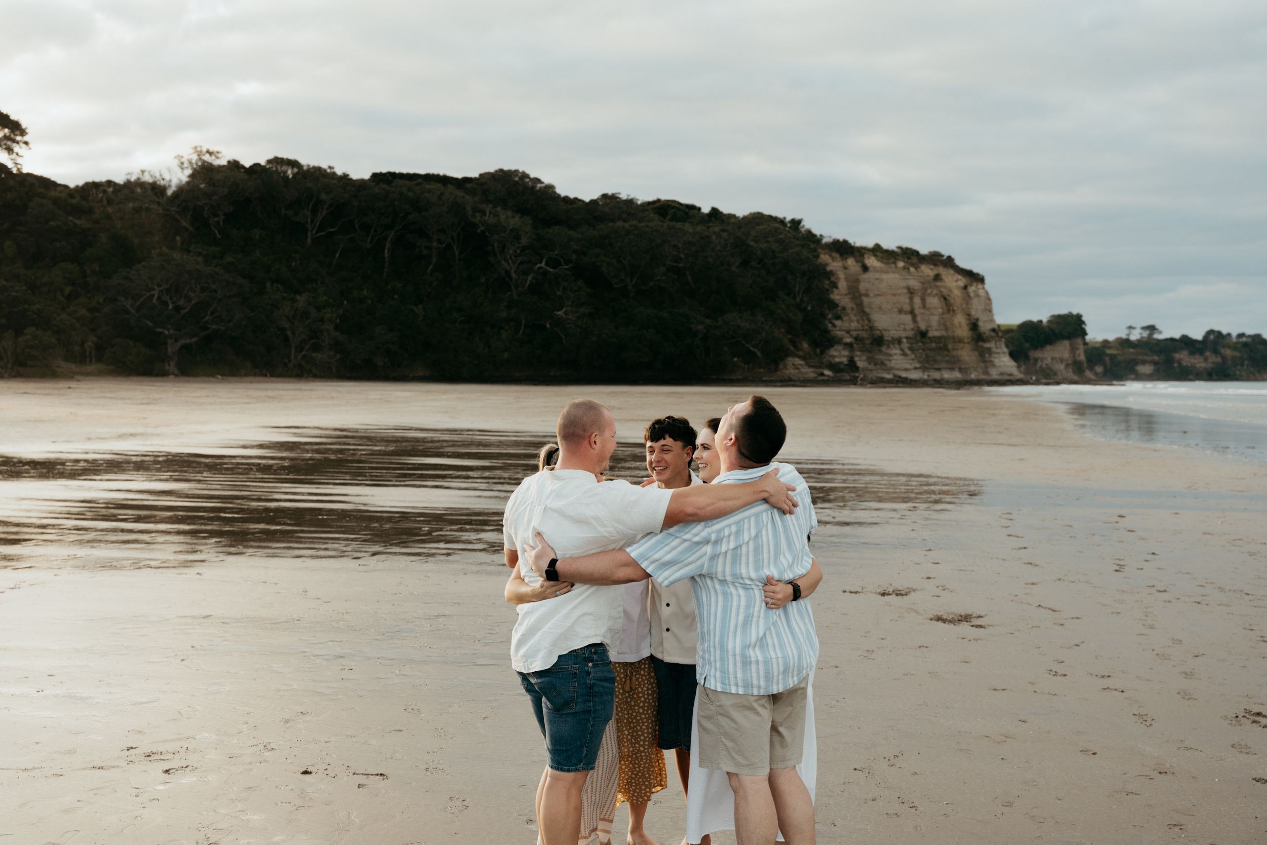 Four friends embracing on a sandy beach with rocky cliffs and lush trees in the background.