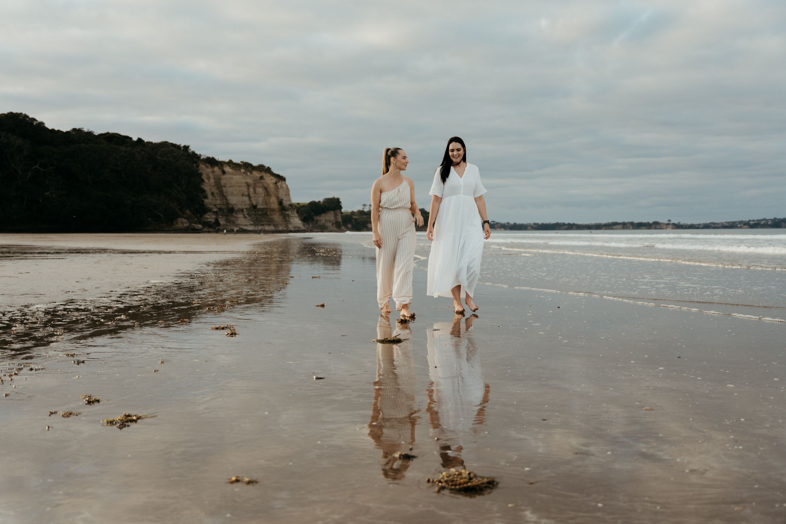 Two brides walk hand-in-hand barefoot on a reflective beach, one in white dress, cliffs in background.