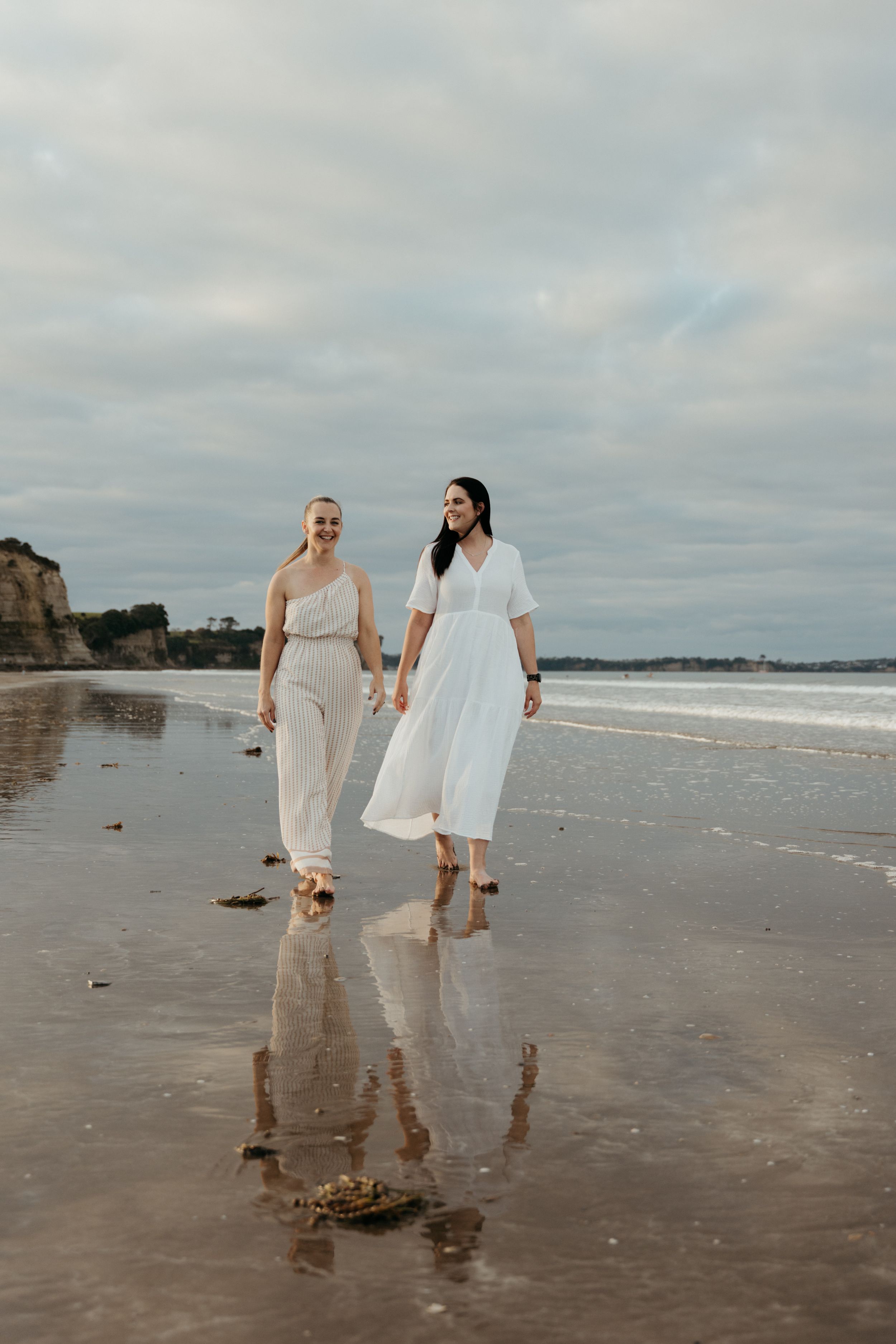 Two women in white dresses walking hand in hand on a reflective beach with cloudy skies.