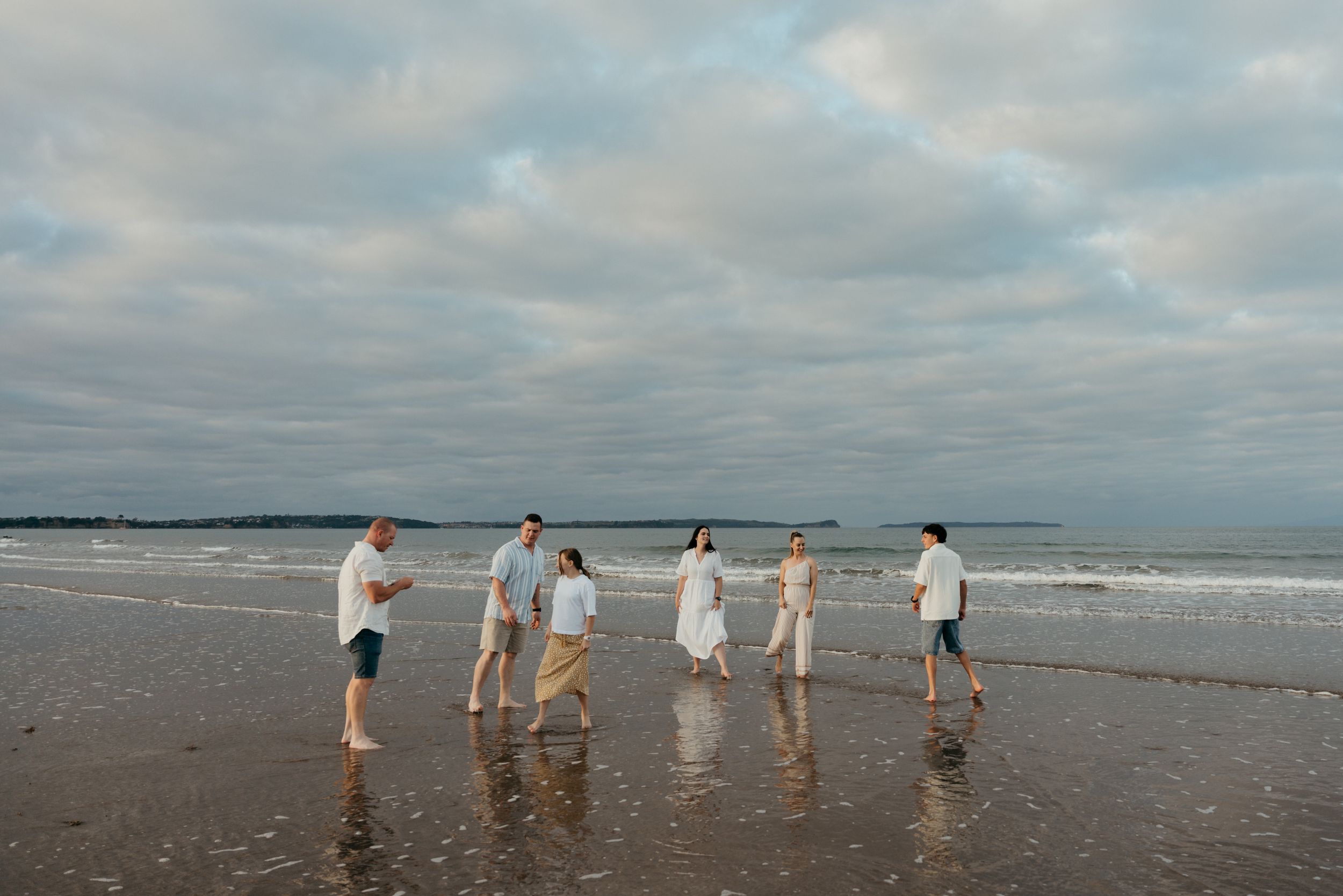 A family of six walks along a reflective beach shoreline under a cloudy sky, dressed in white and blue.