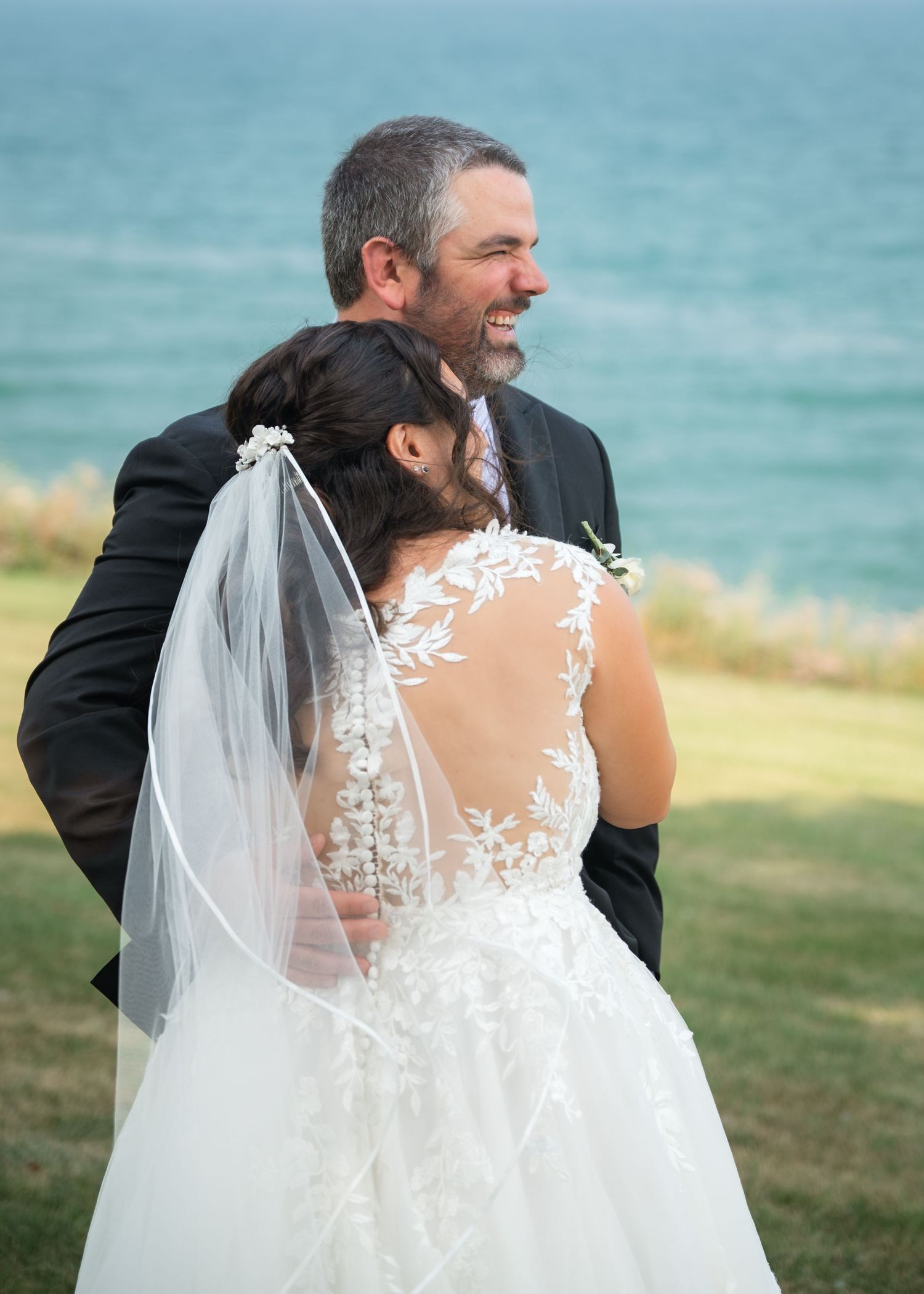 Bride in lace wedding dress with veil embraces groom in black suit outdoors by the ocean.