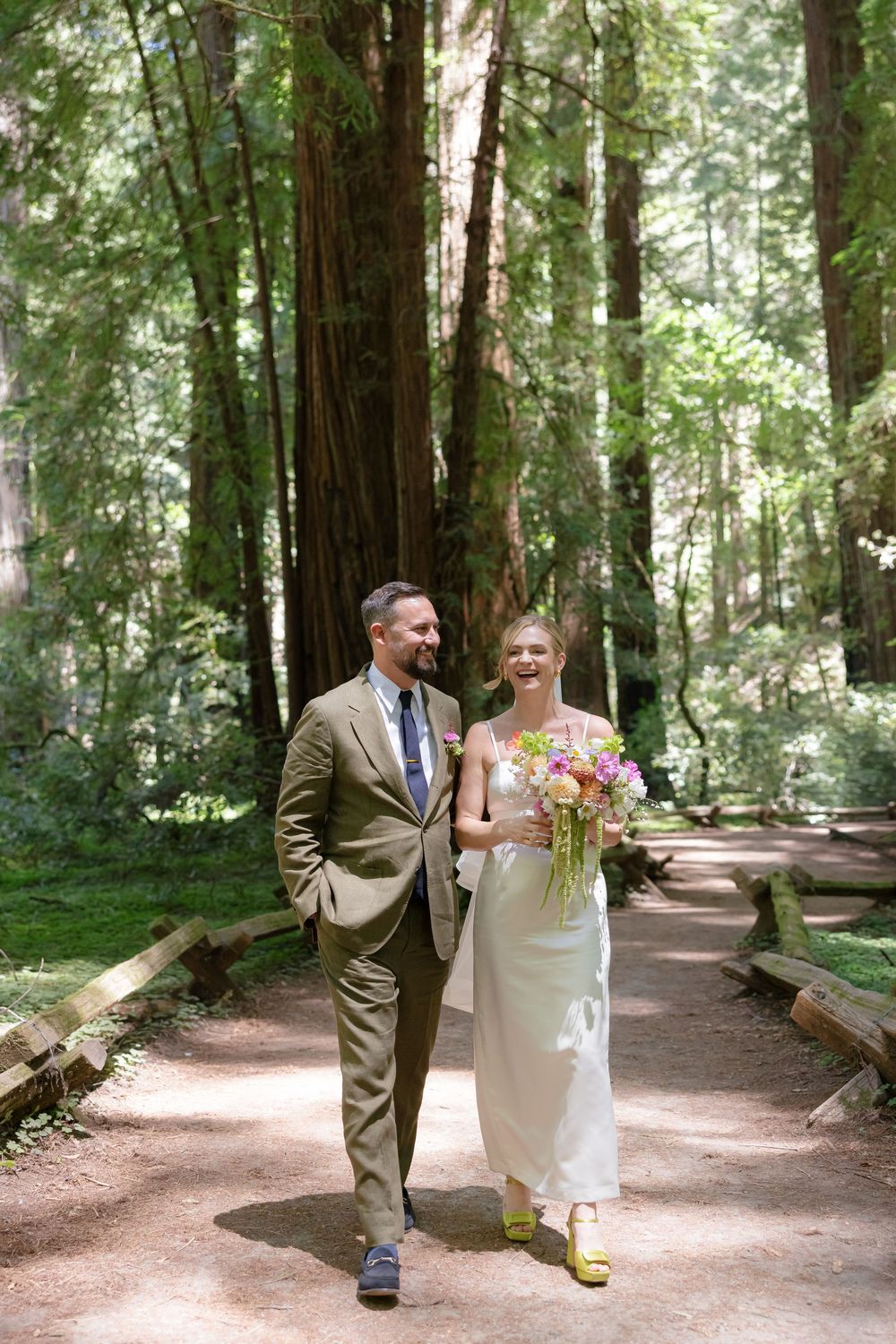 Bride and groom walk hand in hand through a sunlit redwood forest path, bride holding a colorful bouquet.