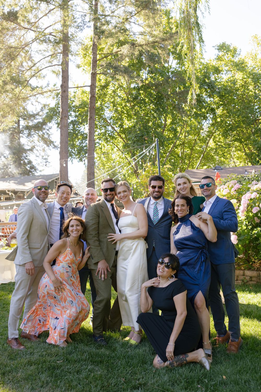 Wedding party group photo on a sunny lawn with tall trees and pink hydrangeas in the background.