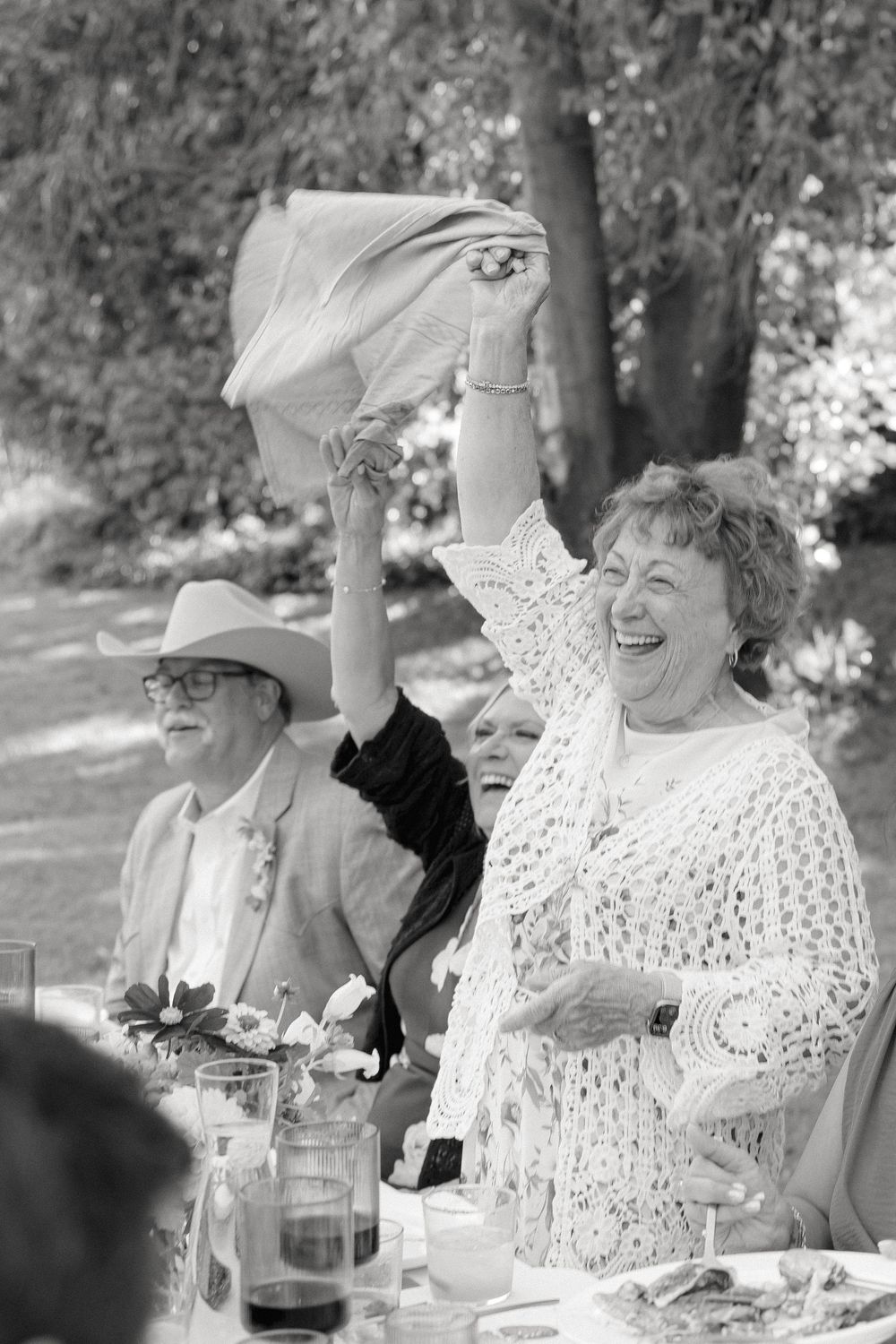 Joyful wedding guest raises napkin and laughs at outdoor garden wedding reception in black and white.