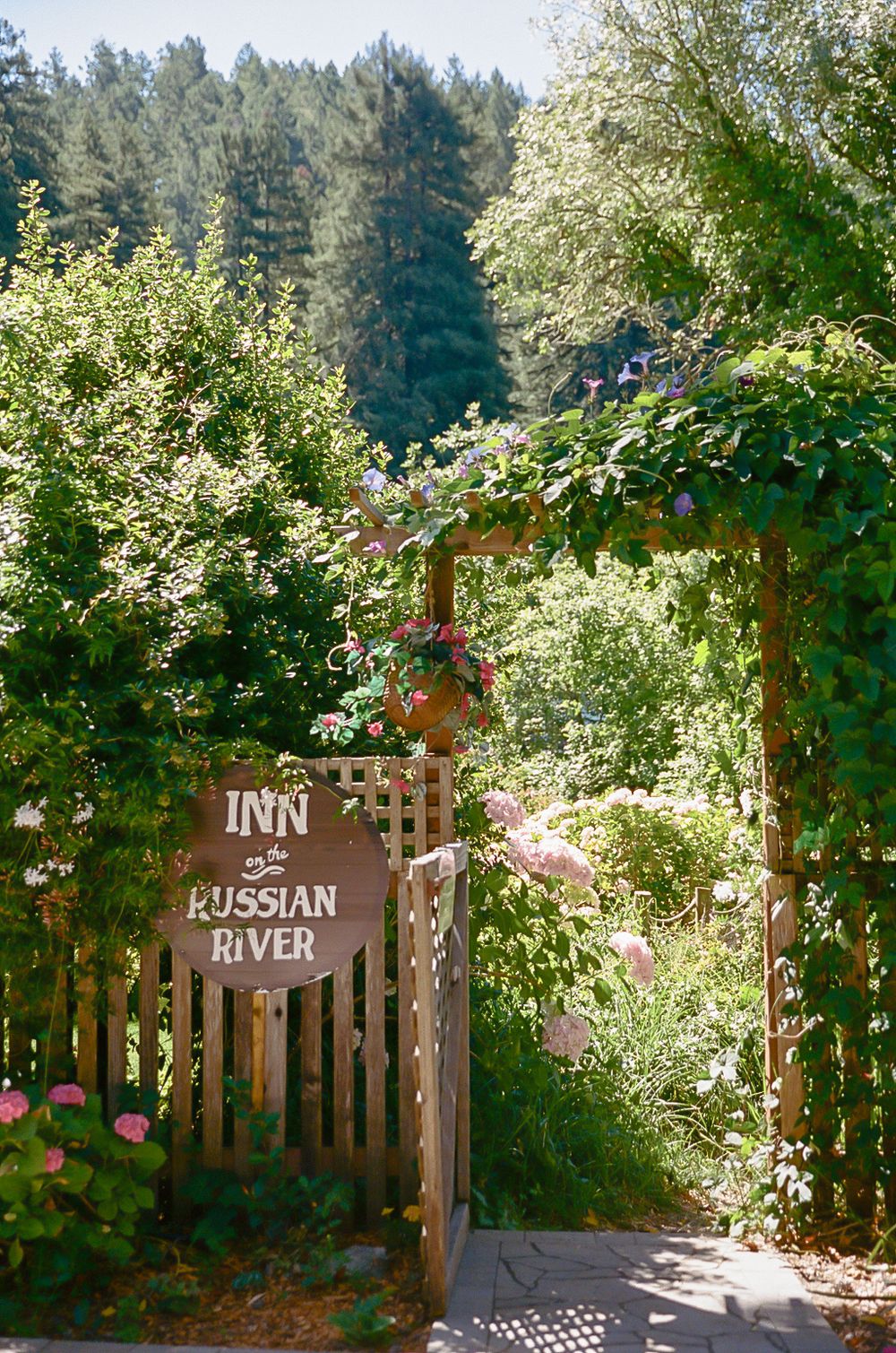 Rustic wooden gate and fence with Inn at Russian River sign surrounded by lush greenery and colorful flowers.