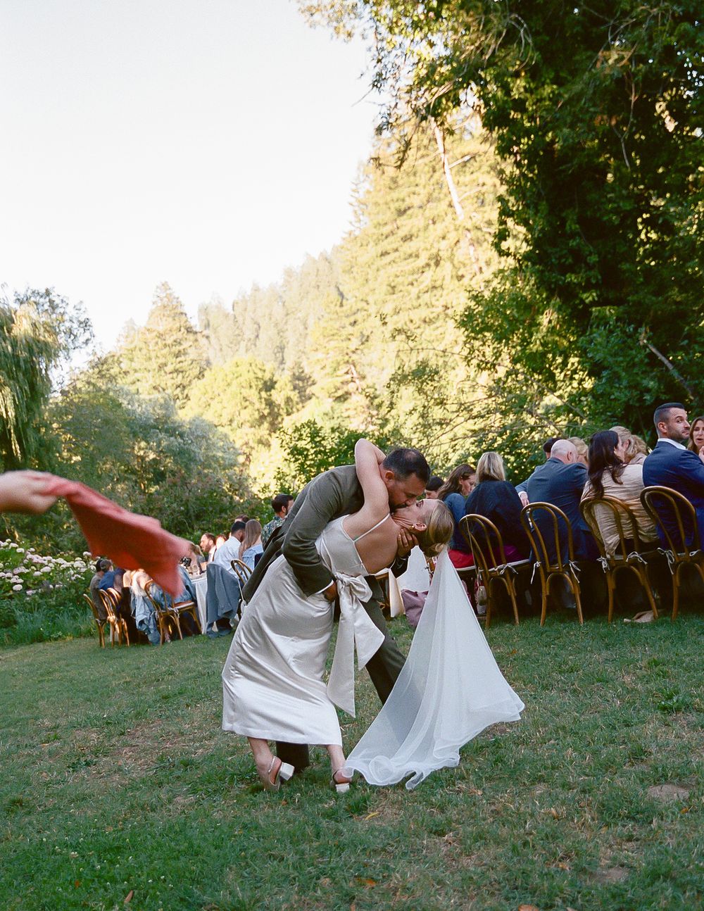 Couple sharing a romantic dip kiss on grass at outdoor wedding reception surrounded by guests and lush trees.