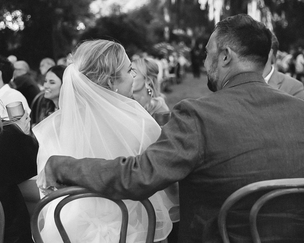 Black and white wedding photo of bride in veil and groom in suit seated together at outdoor ceremony.