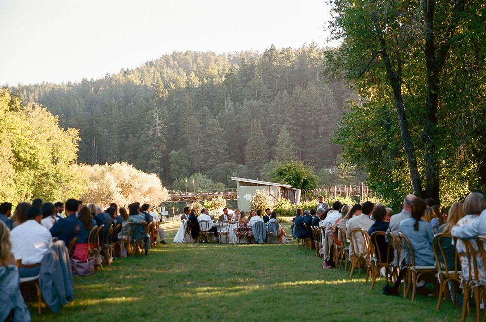 Outdoor wedding ceremony with guests seated on wooden chairs facing altar with forested mountain backdrop.