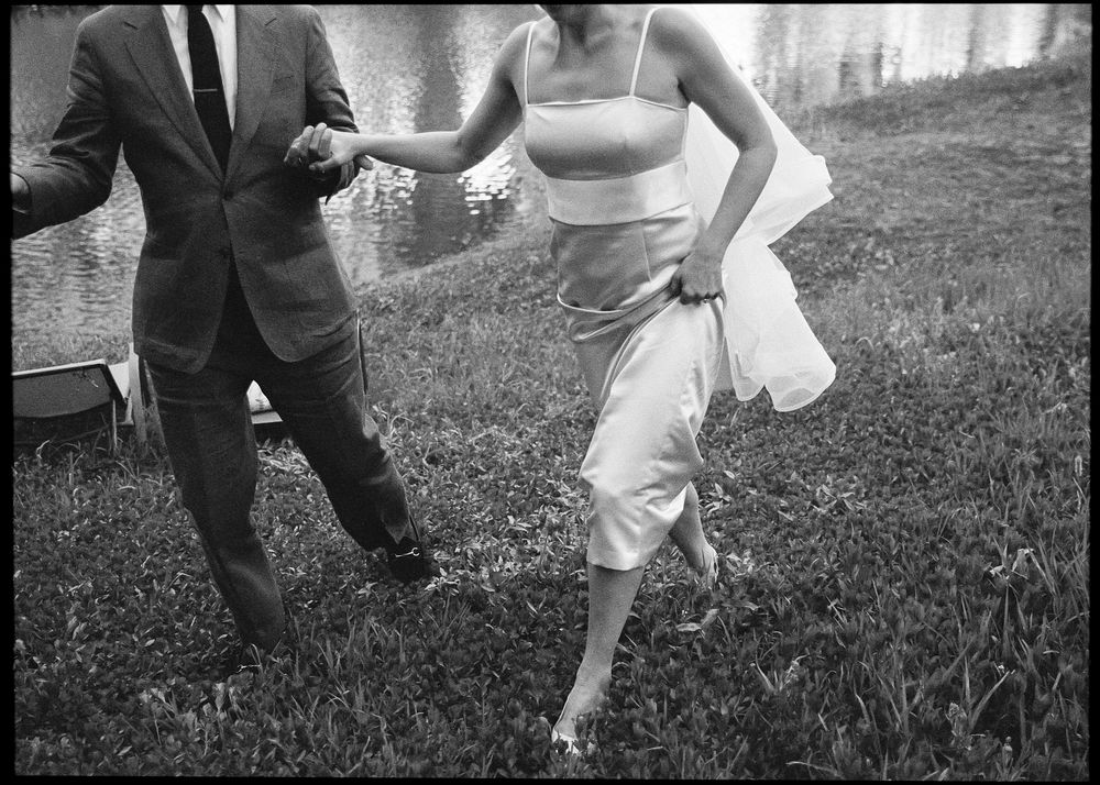 Black and white photo of a couple holding hands while walking on grass near a pond, woman in elegant white dress.