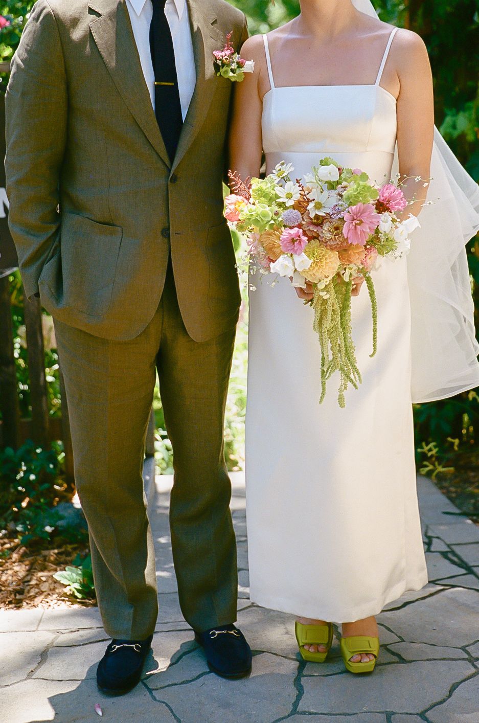 Bride in white slip dress with colorful bouquet stands beside groom in olive suit on stone pathway.