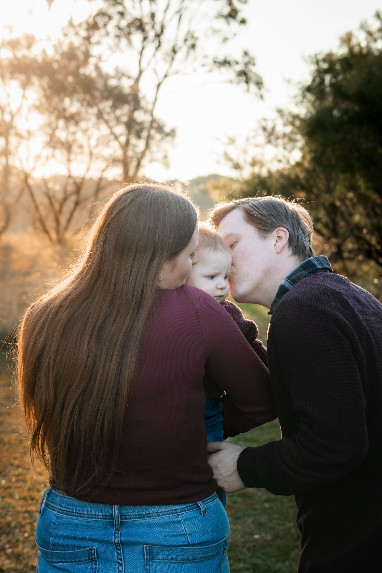 A couple sharing a kiss over their young child outdoors during golden hour sunset in Rouse Hill Regional Park.