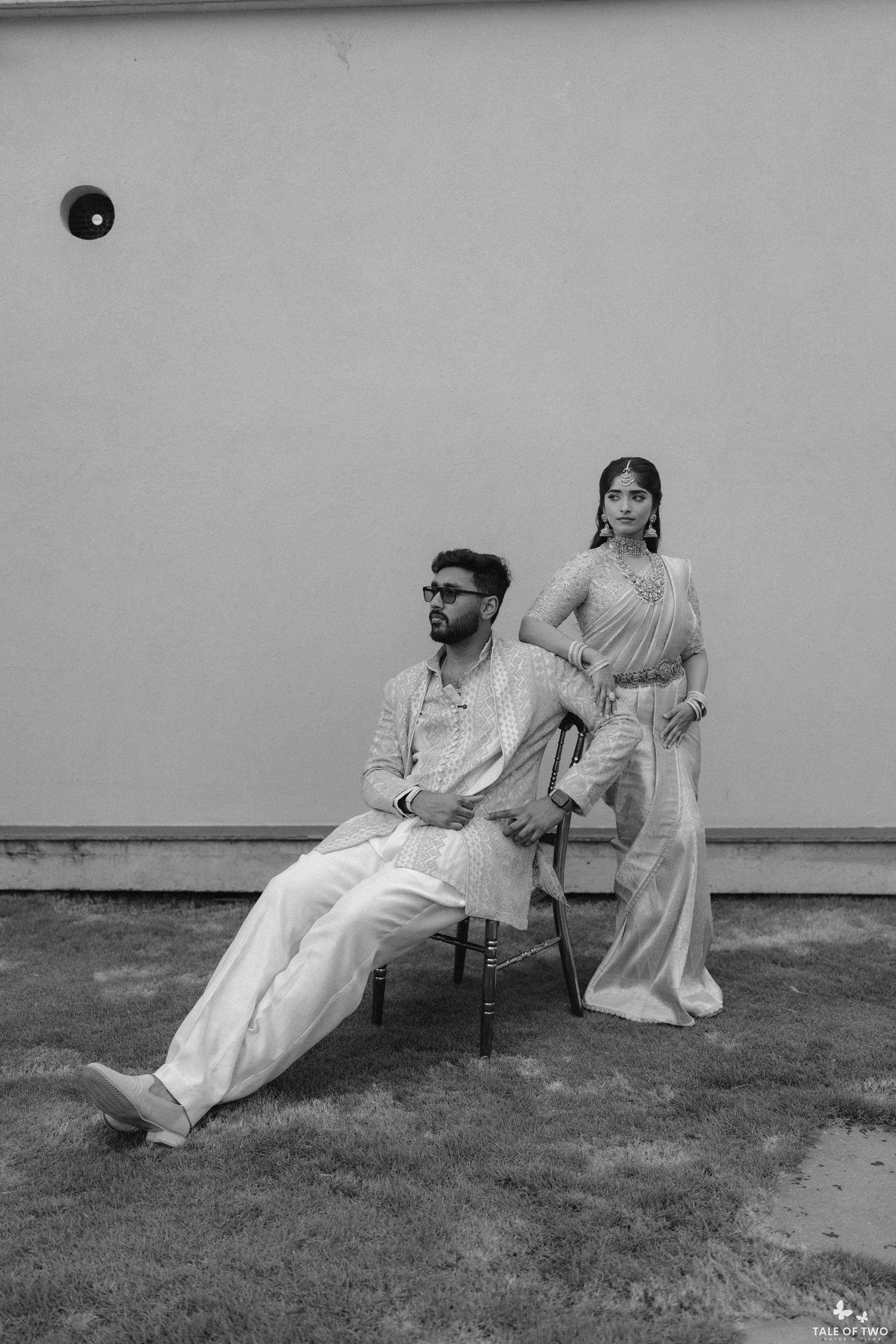 Black and white photo of an Indian couple in traditional attire, man seated in chair, woman standing behind him.