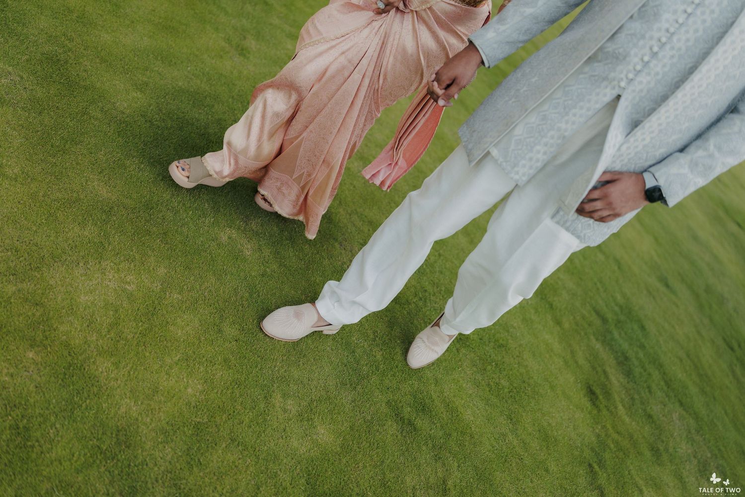 Couple in pastel outfits standing on green grass, wearing white shoes, holding a pink bag.