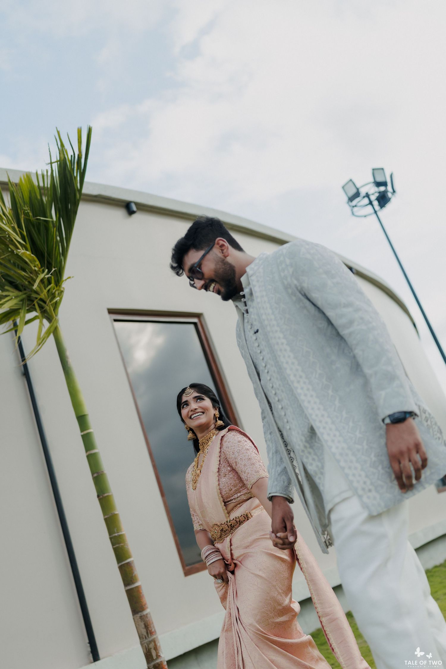 Indian bride in pink saree and groom in grey sherwani holding hands outdoors at a modern venue.