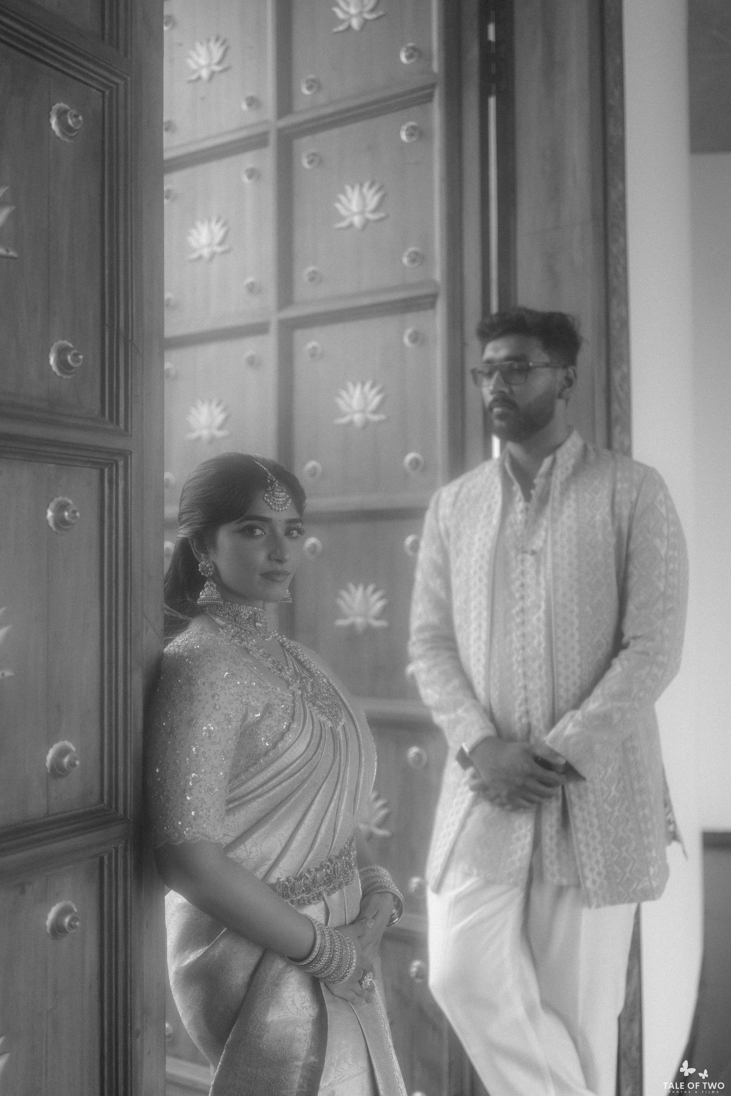 Black and white photo of Indian bride in saree and groom in sherwani posing by ornate wooden door.