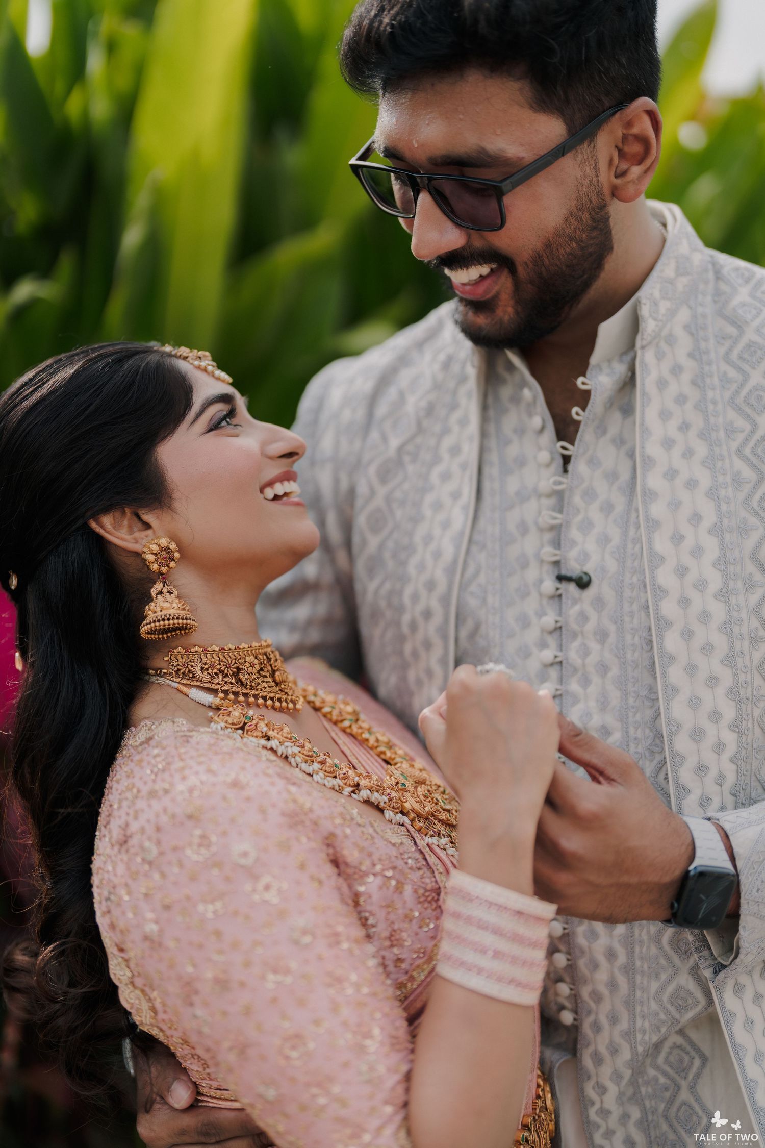 Indian bride in pink lehenga with gold jewelry laughing with groom in white sherwani outdoors.