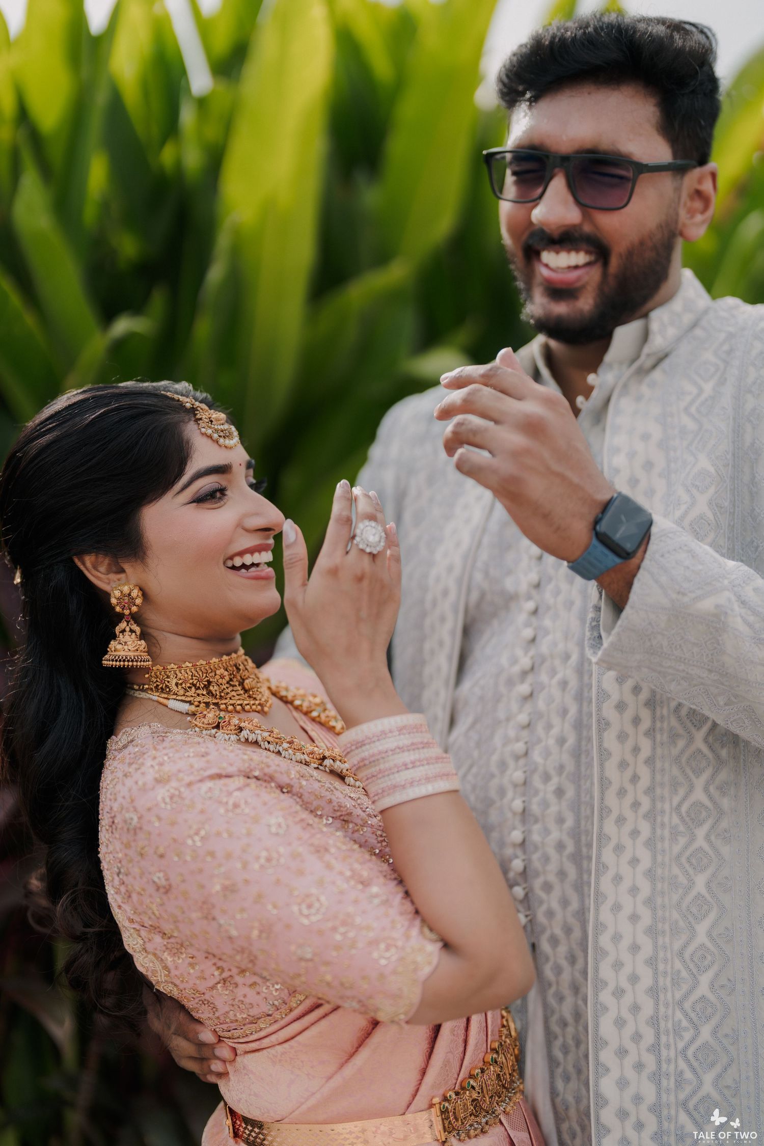 Joyful Indian couple in engagement ceremony, bride in pink lehenga with gold jewelry, groom in grey sherwani.