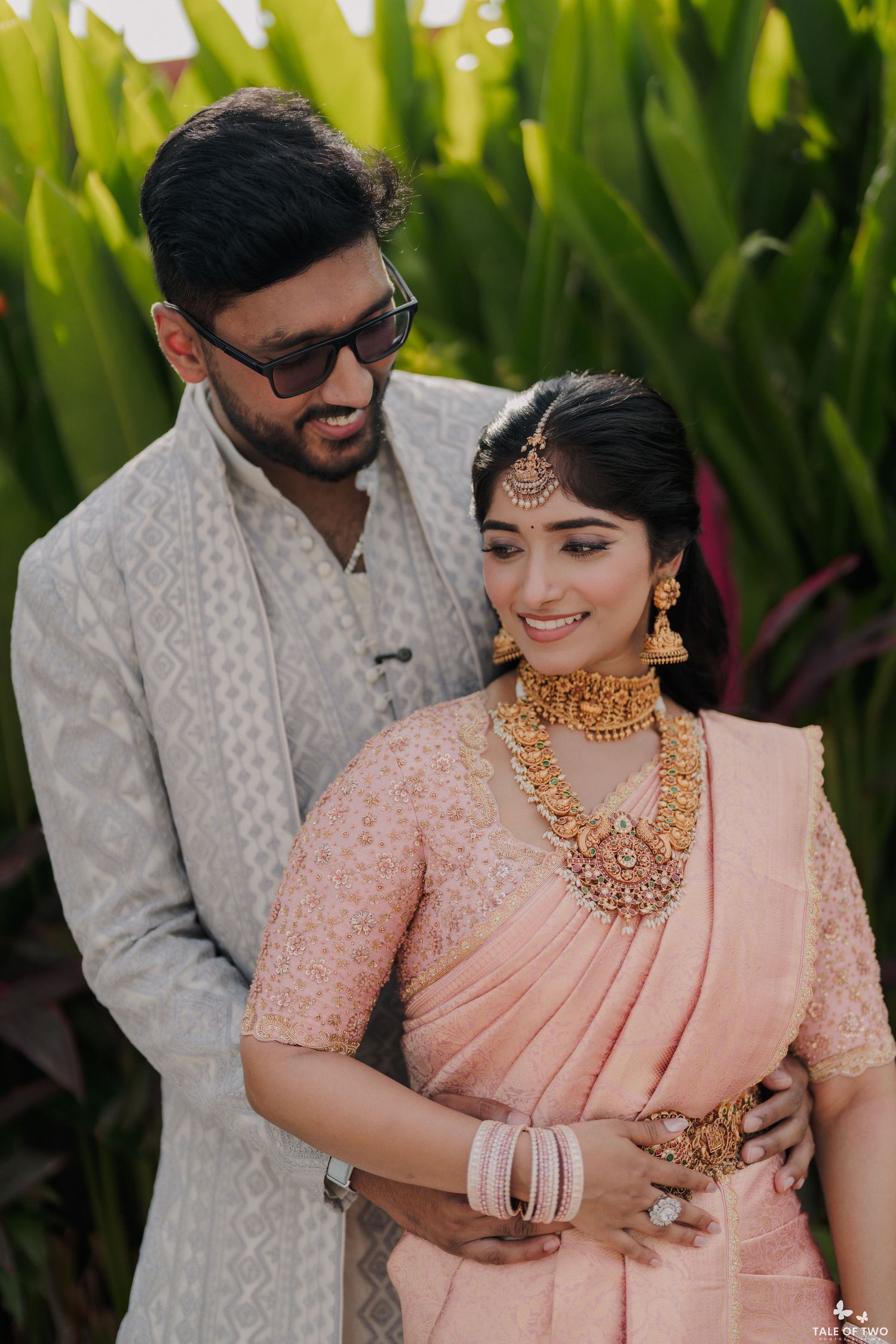 Indian wedding couple embracing outdoors, bride wearing pink silk saree with elaborate gold temple jewelry and groom in grey sherwani.