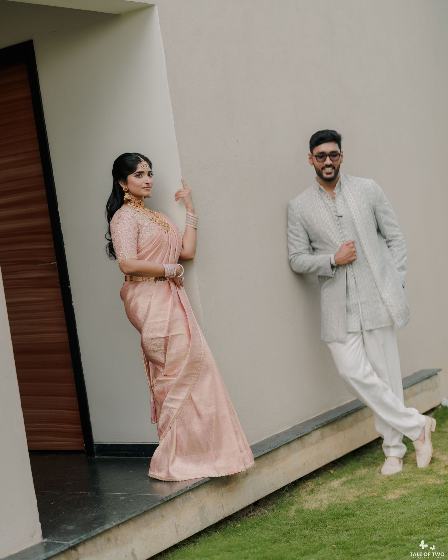 Elegant Indian couple posing outdoors, bride in blush pink saree and groom in grey Indo-western sherwani suit.