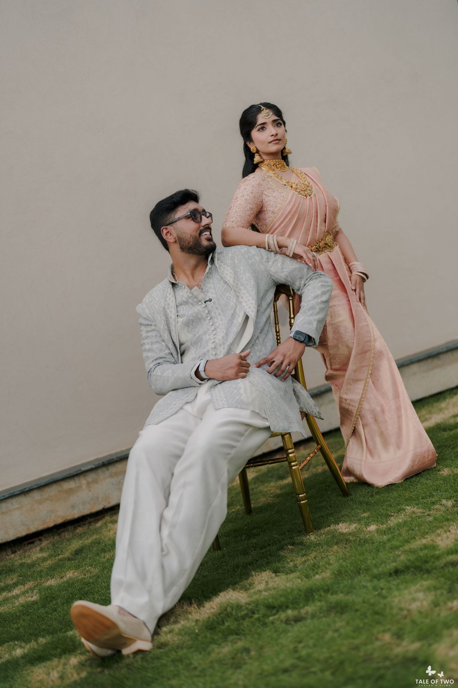Indian couple in traditional attire, groom in white sherwani seated, bride in pink saree standing behind.