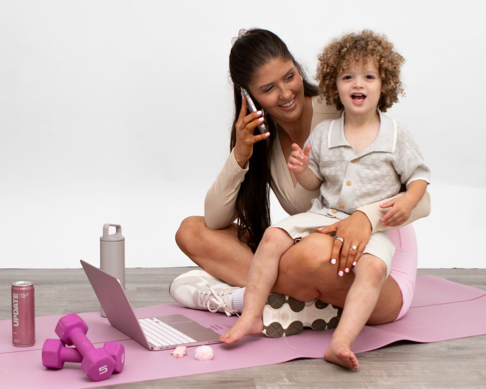 Mom on pink yoga mat talks on phone with curly-haired toddler, laptop and dumbbells nearby.