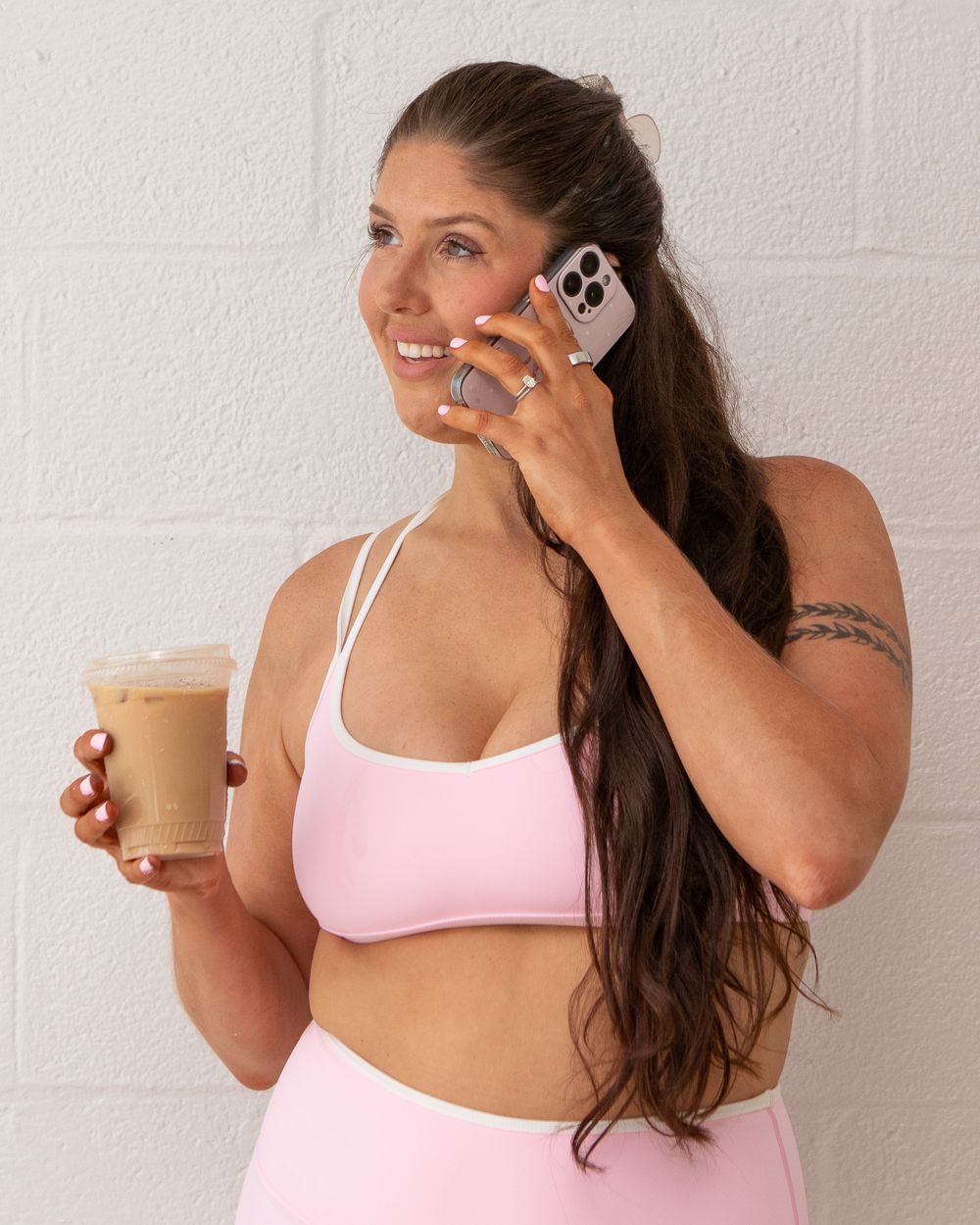Woman in pink sports bra holds iced coffee and talks on phone, smiling, with long dark hair and tattoo.