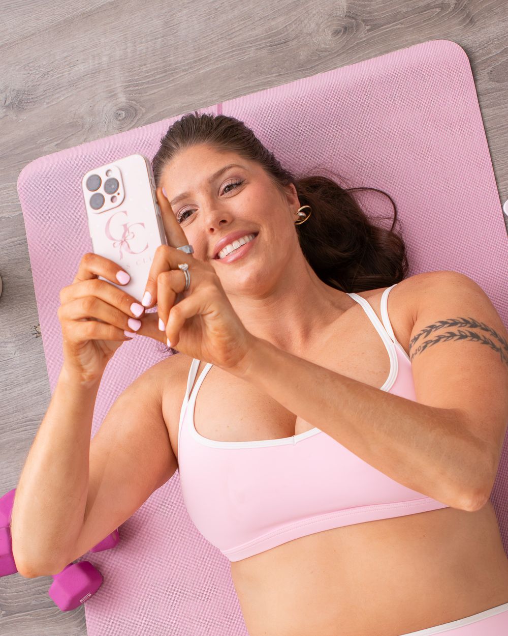 Woman in pink sports bra lying on yoga mat, smiling while holding a smartphone during a workout break.