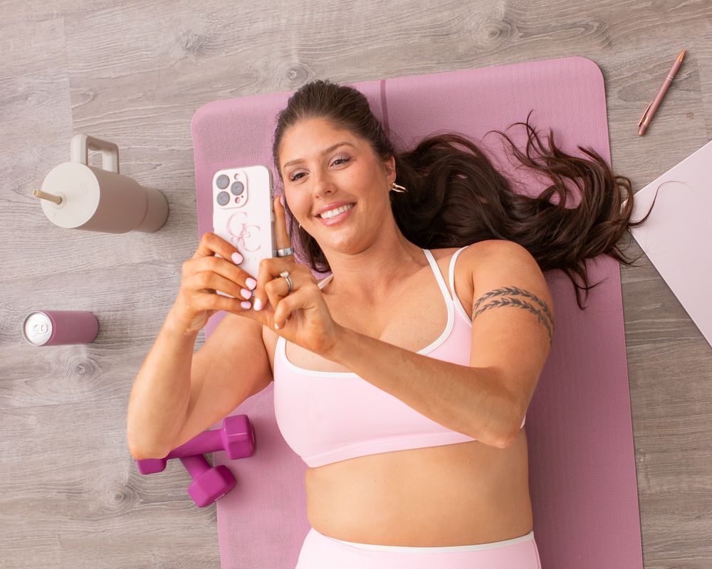 Woman in pink sports bra lying on yoga mat taking selfie with phone, surrounded by dumbbells and workout gear.