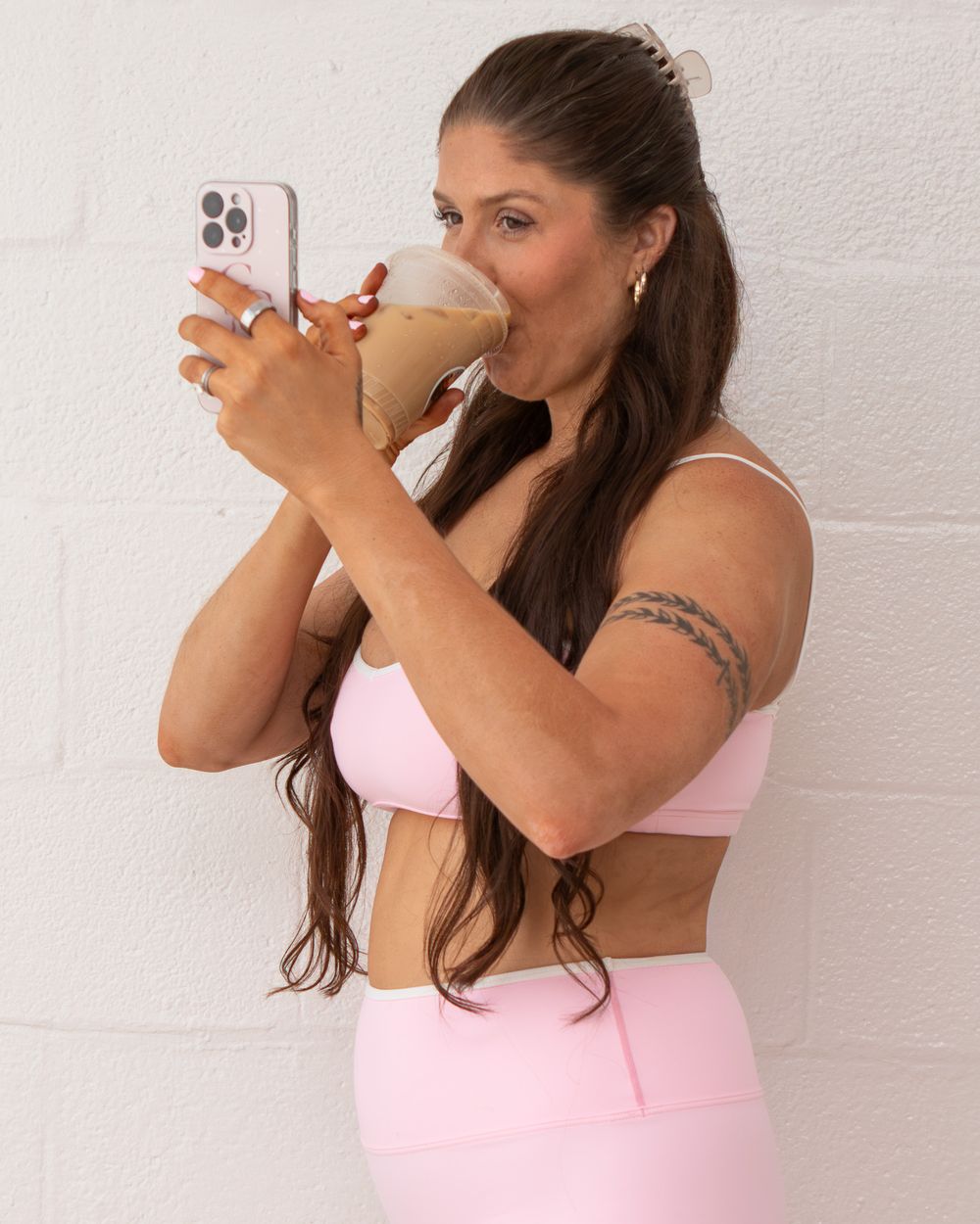 Woman in pink sports bra and leggings drinking a smoothie while holding a phone against a white wall.