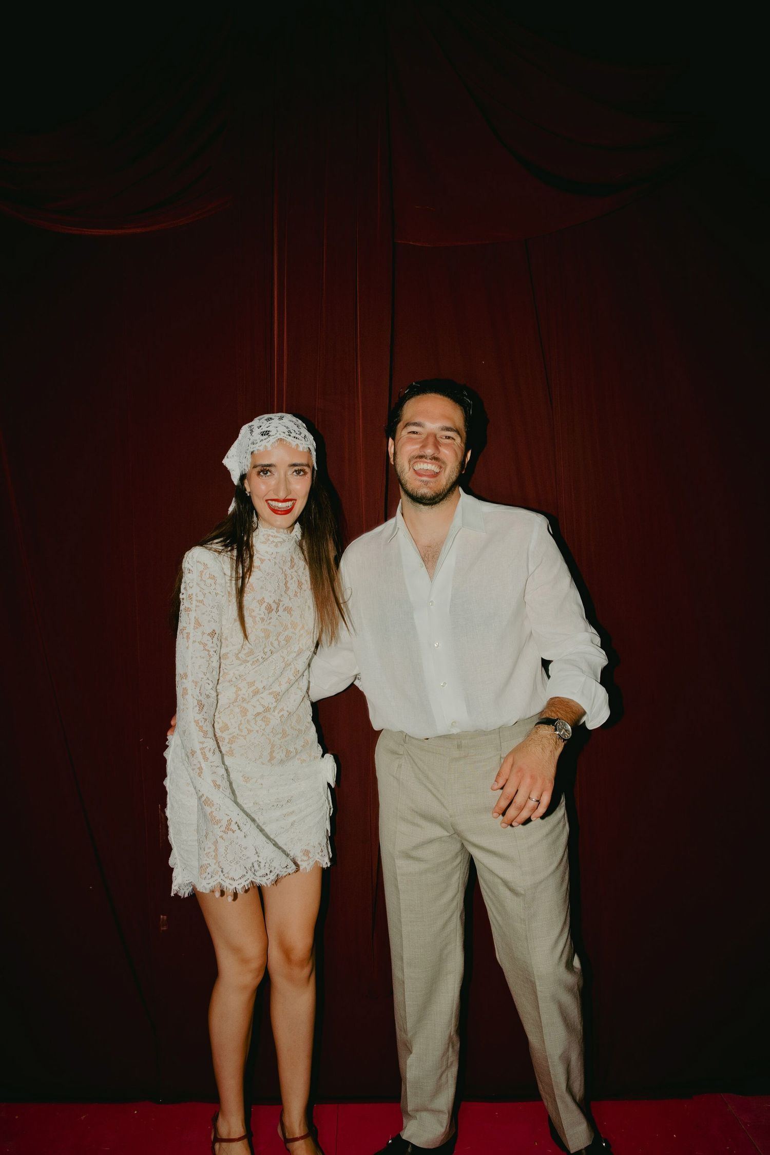 A smiling couple in white outfits pose together against a dark red curtain backdrop.