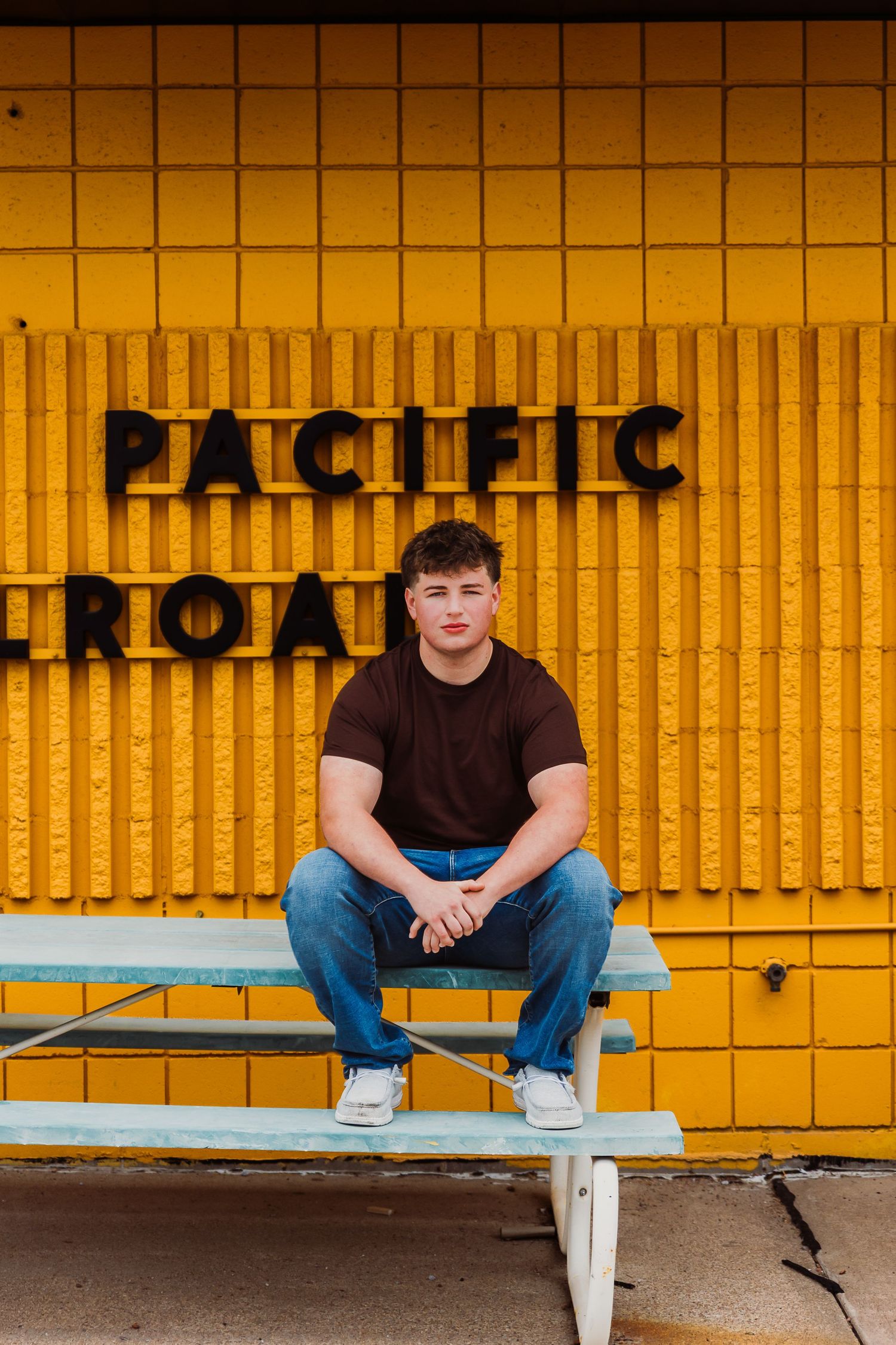 Young man in brown shirt and jeans leaning against a blue brick wall with a large industrial window.