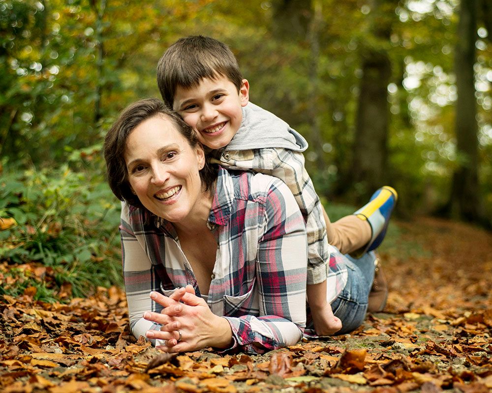 Smiling mother and young son lie playfully on autumn leaves in a sunlit forest; he hugs her from behind, conveying warmth and joy.