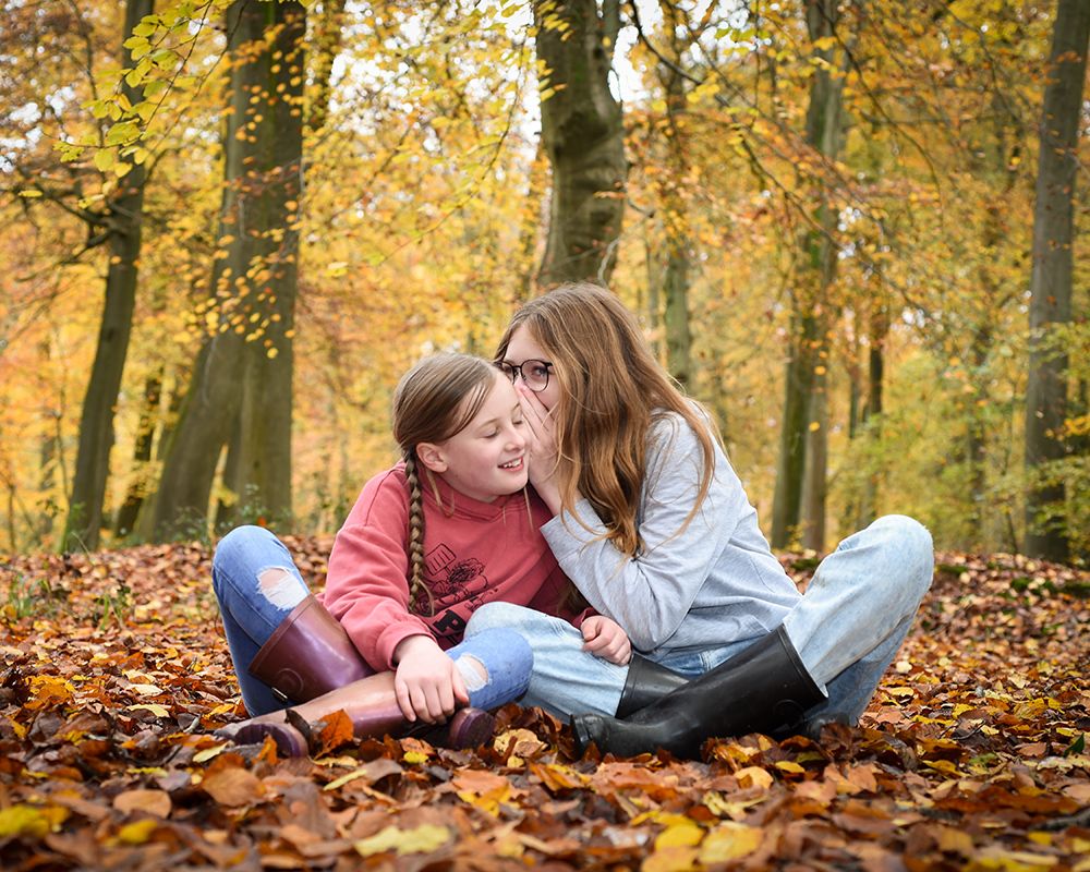 In an autumn forest, a smiling mother kisses her laughing daughter’s cheek as they sit together on colorful fallen leaves.