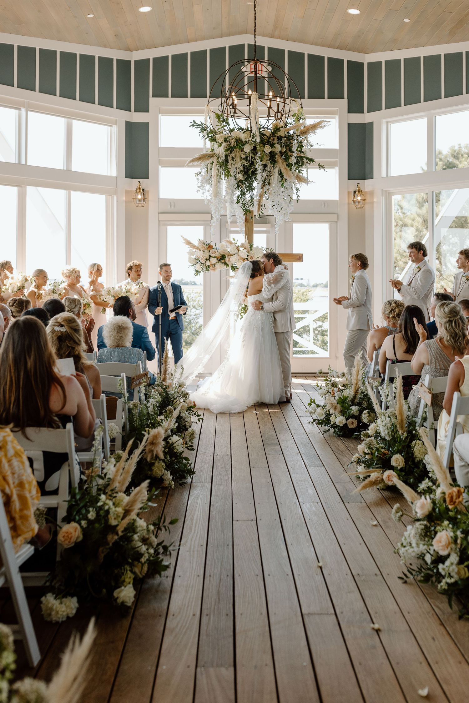 Bride and groom share first kiss at elegant indoor wedding ceremony with floral aisle decorations and large windows.