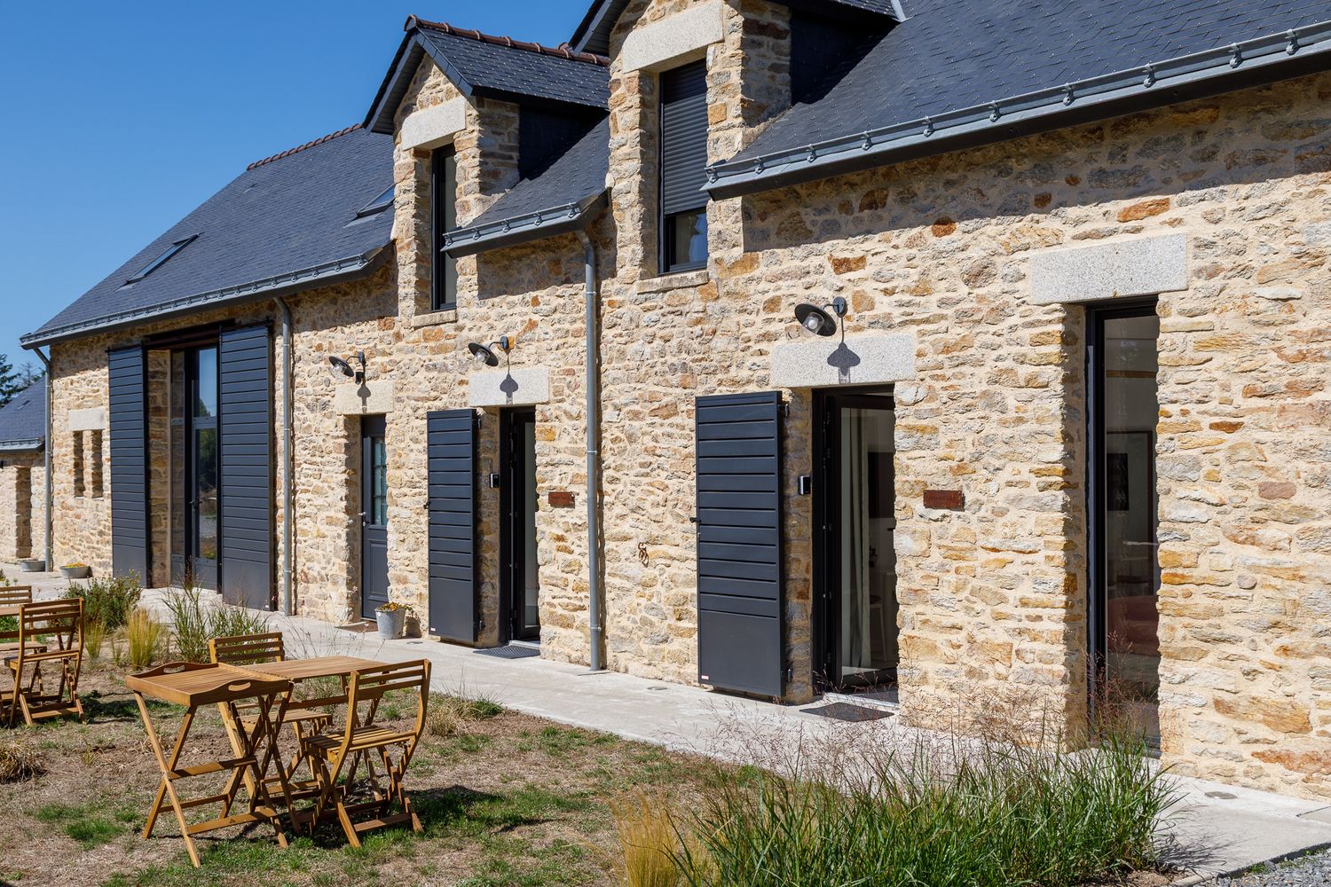 Chaumière en pierre française rénovée, avec volets sombres, mobilier d'extérieur en bois et toit en ardoise, par temps ensoleillé.