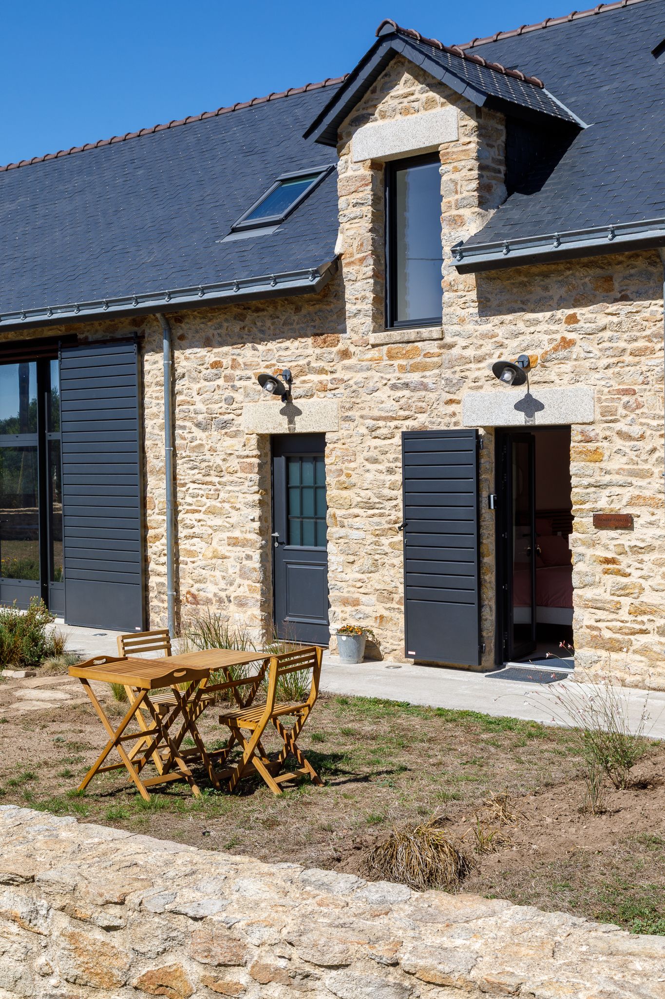 Extérieur d'une ferme en pierre avec volets gris foncé, fenêtre de toit, toit en ardoise et un ensemble table et chaises de bistrot en bois