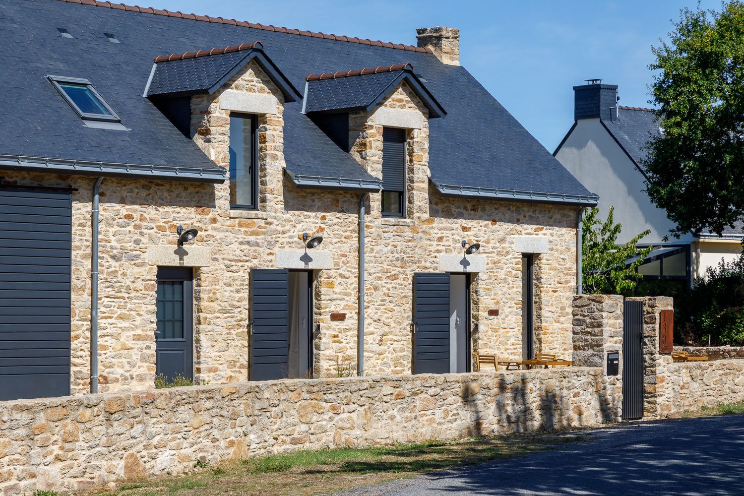 Maison bretonne traditionnelle en pierre avec volets gris, toit en ardoise et muret de clôture en pierre, située dans une rue calme.