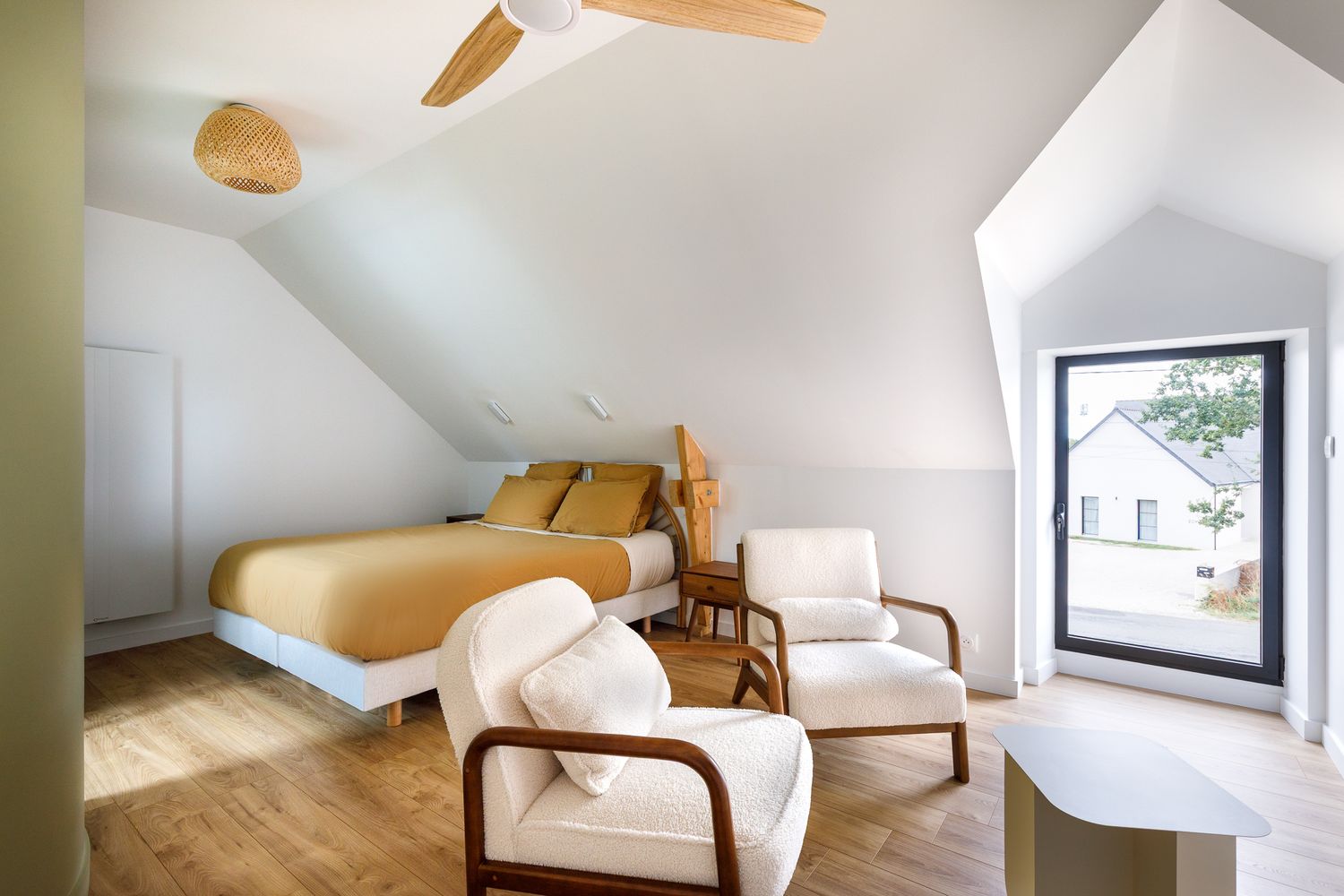 Chambre lumineuse sous les combles avec une parure de lit moutarde, des fauteuils blancs en tissu bouclé, un parquet en bois