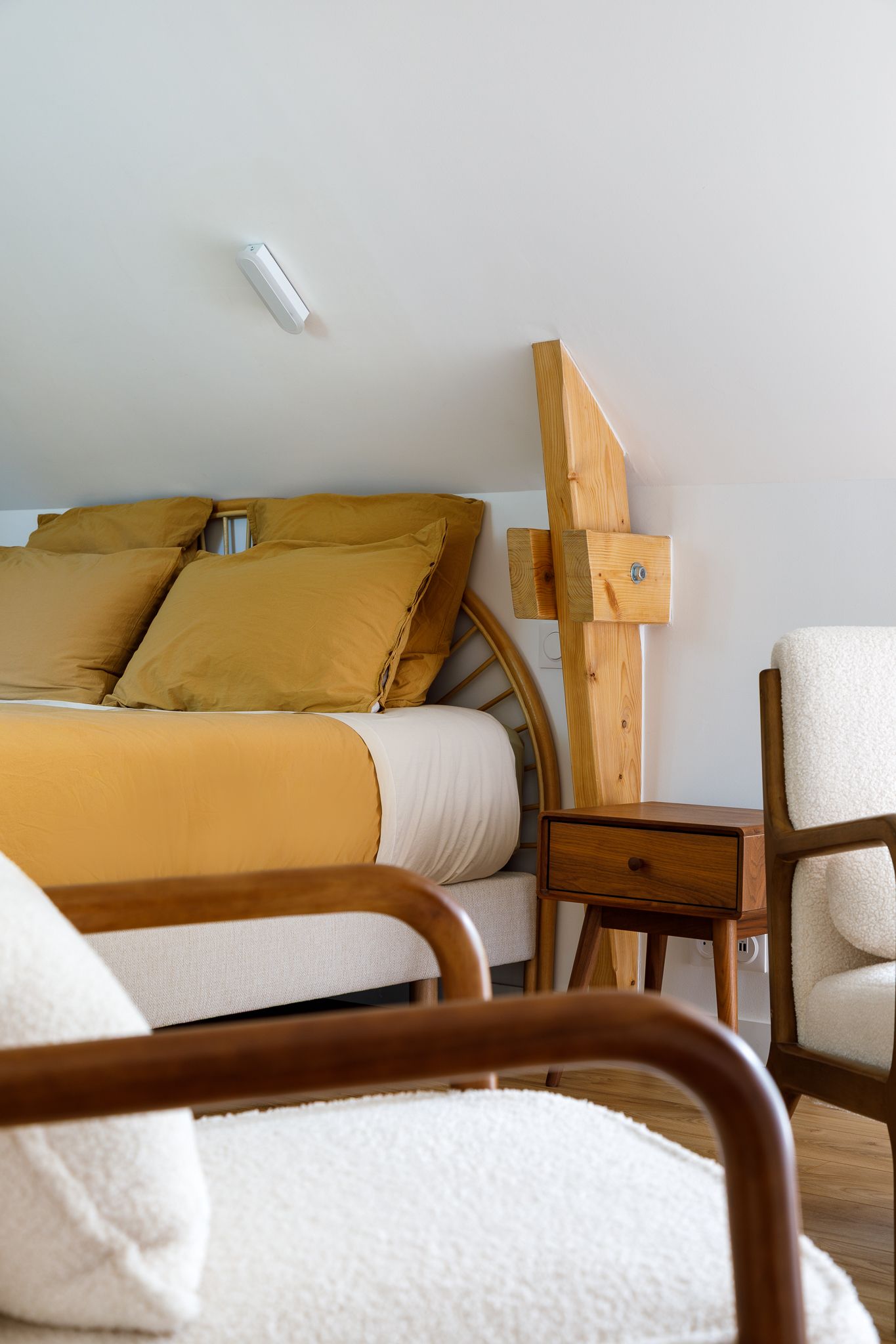 Gros plan sur une chambre sous les combles avec une parure de lit jaune moutarde, une tête de lit en bois, un fauteuil en tissu bouclé