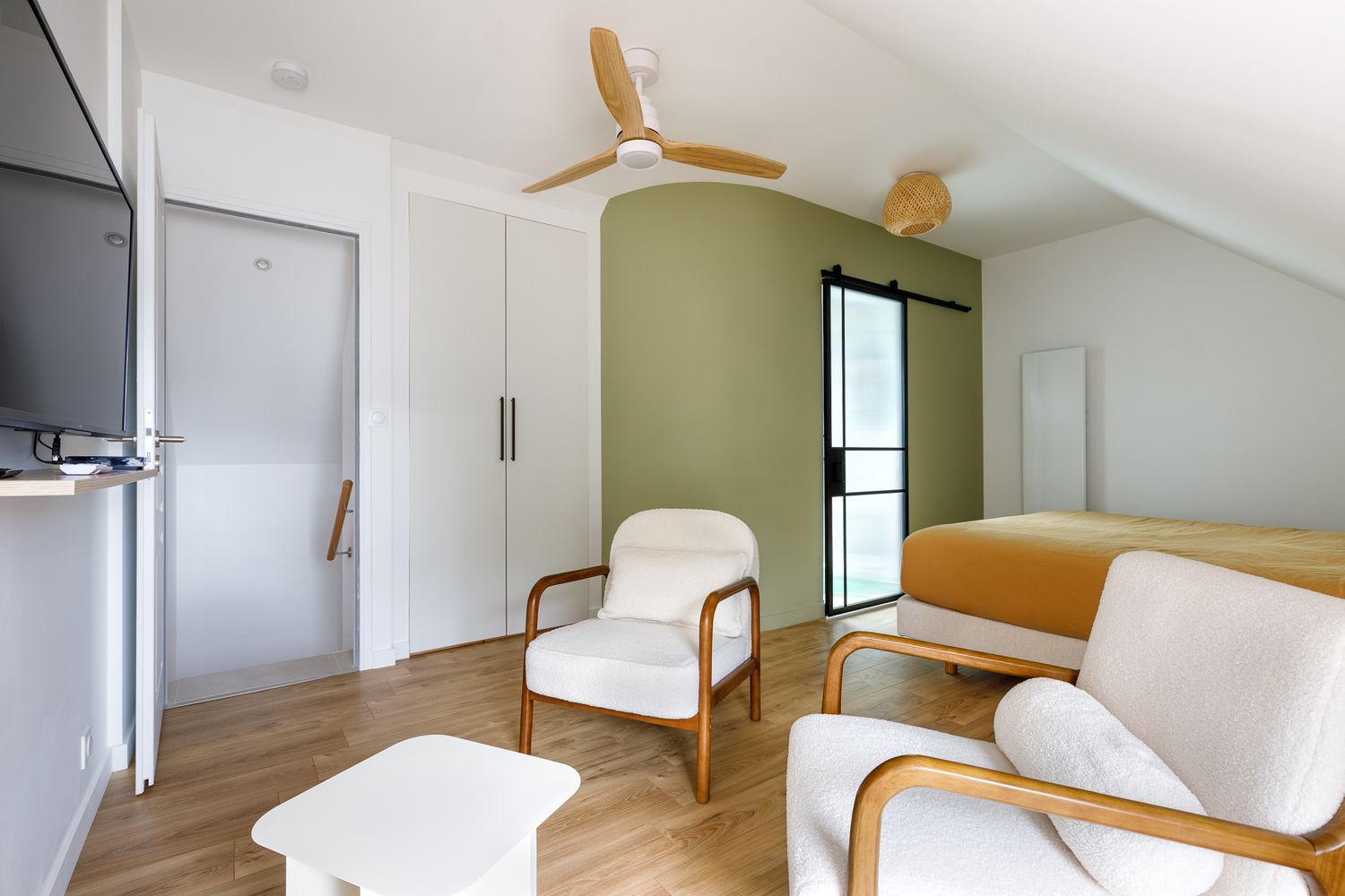 Salon moderne et lumineux avec un mur d'accent vert olive, des fauteuils blancs, un ventilateur de plafond en bois et du parquet.